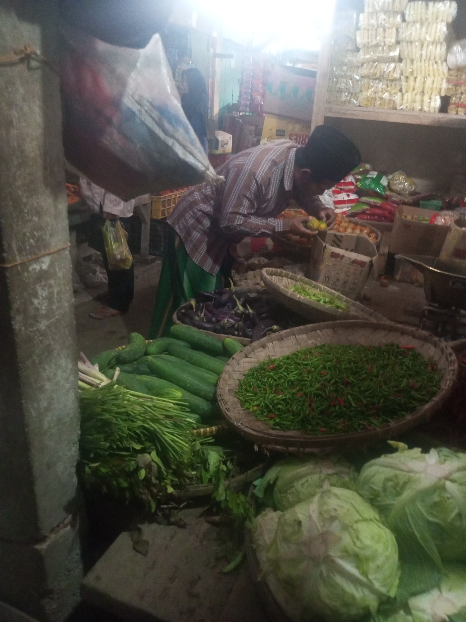 The image depicts a market scene in Kalibeber, Indonesia. In the foreground, a large concrete pillar is visible on the left. To the right of the pillar, a man wearing a plaid shirt and a black peci (cap) is bending over a basket filled with small green and red chilies. Behind him, various vegetables are displayed, including long green beans, cucumbers, and eggplants. Further in the background, shelves are stocked with packaged goods, and other market stalls are partially visible. The lighting is dim, suggesting an indoor or shaded area.