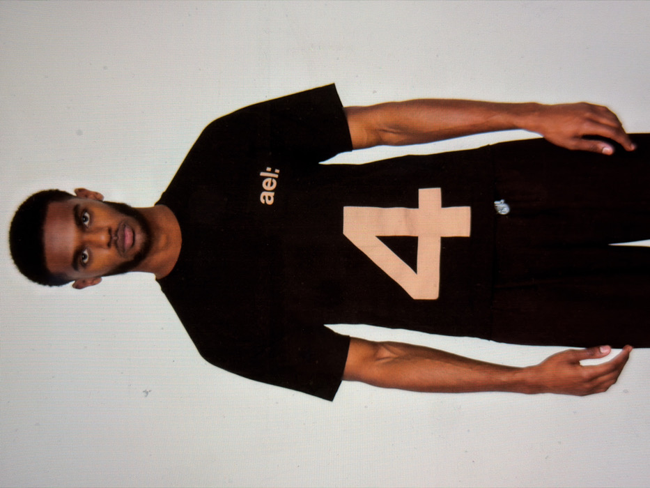 A single male individual is depicted in a full-body, front-facing studio portrait. The subject has dark skin, short curly black hair, and a trimmed beard and mustache. He wears a black short-sleeved crew-neck t-shirt featuring the light beige text "ael:" on the upper right chest and a large light beige numeral "4" prominently displayed below it. He is also dressed in black trousers, exhibiting a light-colored metal or plastic drawstring aglet visible on the right side of the waistband. His arms hang relaxed at his sides, and his gaze is directed straight at the viewer with a neutral expression. The background is a plain, smooth light grey surface, characteristic of a photography studio. No specific location within the city of London, United Kingdom, is discernable from the visual elements present in the image.