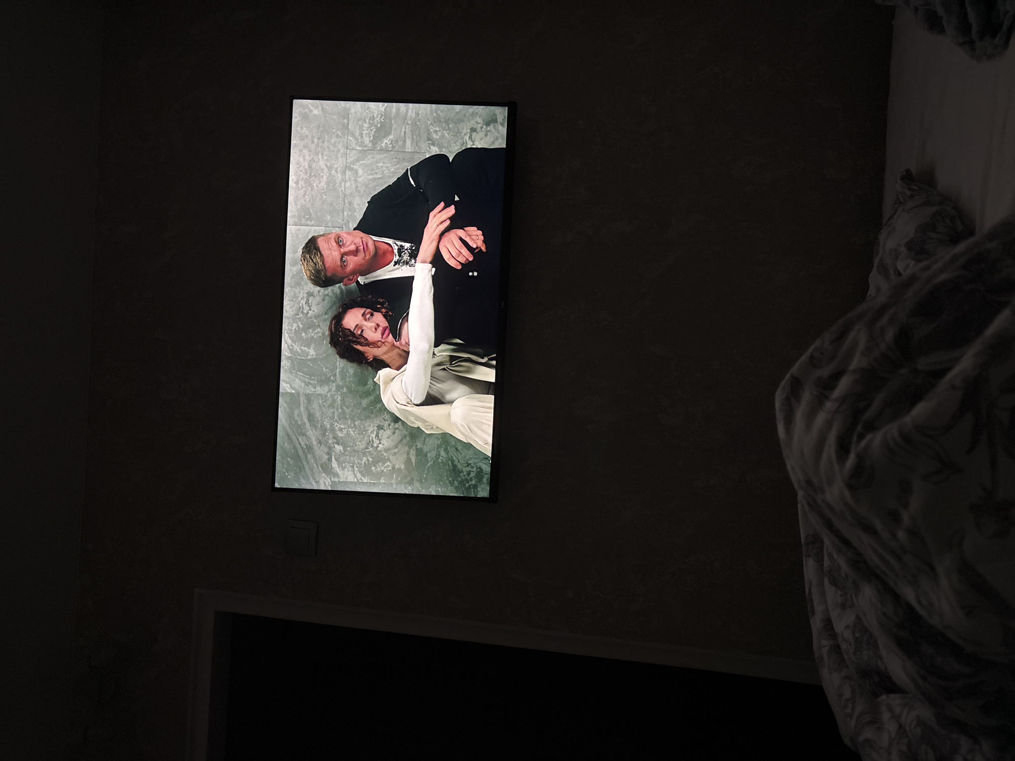 A television displays a scene of a man and a woman sitting closely on a textured, grey background. The man is on the right, looking forward with a serious expression, wearing a black jacket over a white shirt. The woman is on the left, leaning her head on the man's shoulder, her eyes closed and her hair dark and curly. She is wearing a light-colored outfit. The television is mounted on a dark, textured wall. In the foreground, a bed is visible, covered with a patterned duvet. The scene is dimly lit, suggesting it is nighttime or the room is dark.