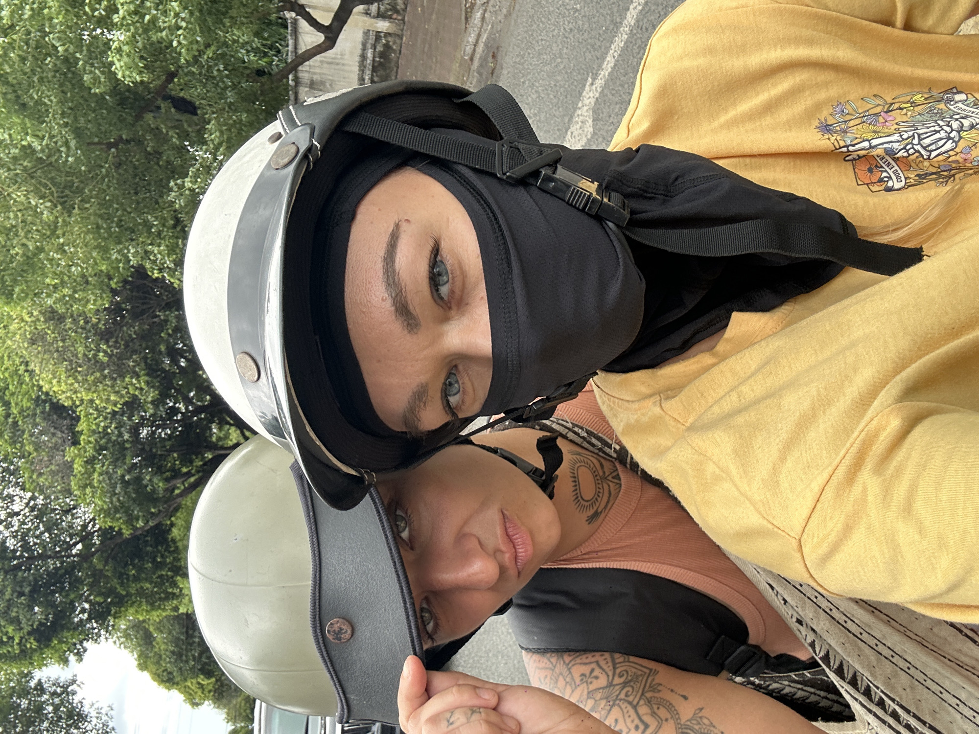 Two fair-skinned women pose closely for a selfie, both wearing helmets and riding a motorcycle. The woman in the back wears a white and gray helmet with a black balaclava covering most of her face. She is wearing a yellow sweatshirt with a graphic design on the chest. The woman in the front wears a light green helmet and has tattoos on her arm, neck, and hand. The environment appears to be an urban street with green trees in the background. The location is Đồng Nai, Vietnam.