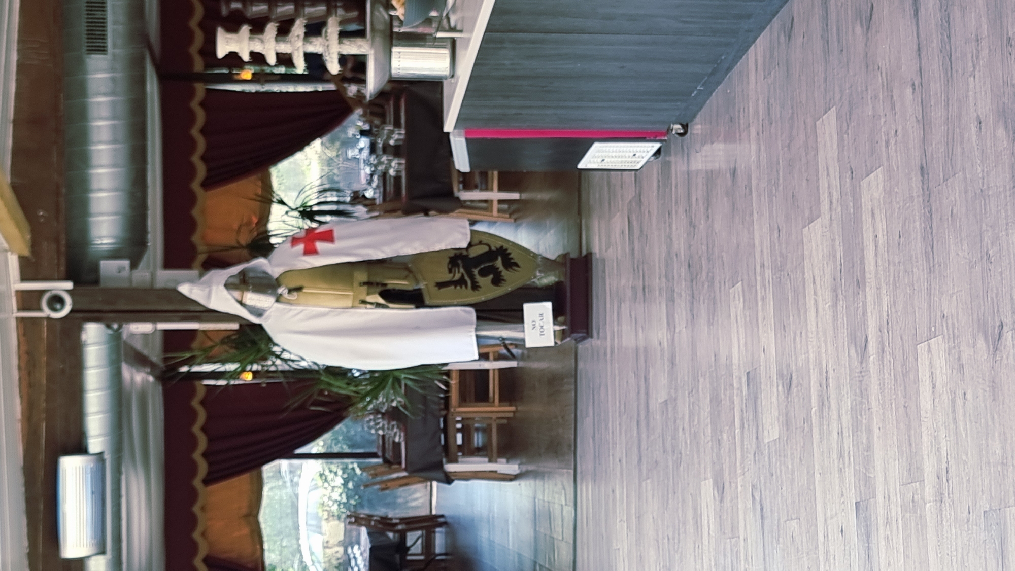The image depicts the interior of a restaurant, possibly in Besalú, Spain, as suggested by the medieval-themed decor. In the foreground, a mannequin dressed as a medieval knight stands prominently, featuring a white hooded cape over golden armor and a shield bearing a black lion emblem and a red cross. A small sign on a stand in front of the mannequin reads "NO TOCAR" (Do Not Touch).
The background reveals a dining area with tables and chairs set for patrons. Drapes in shades of red and gold hang from the windows, adding to the restaurant's ambiance. Various elements of the restaurant's infrastructure are visible, including air conditioning units, lighting fixtures, and what appears to be a service counter with a refrigerator unit. The flooring is a light-colored wood-effect laminate. The overall scene suggests a restaurant with a historical or thematic design.