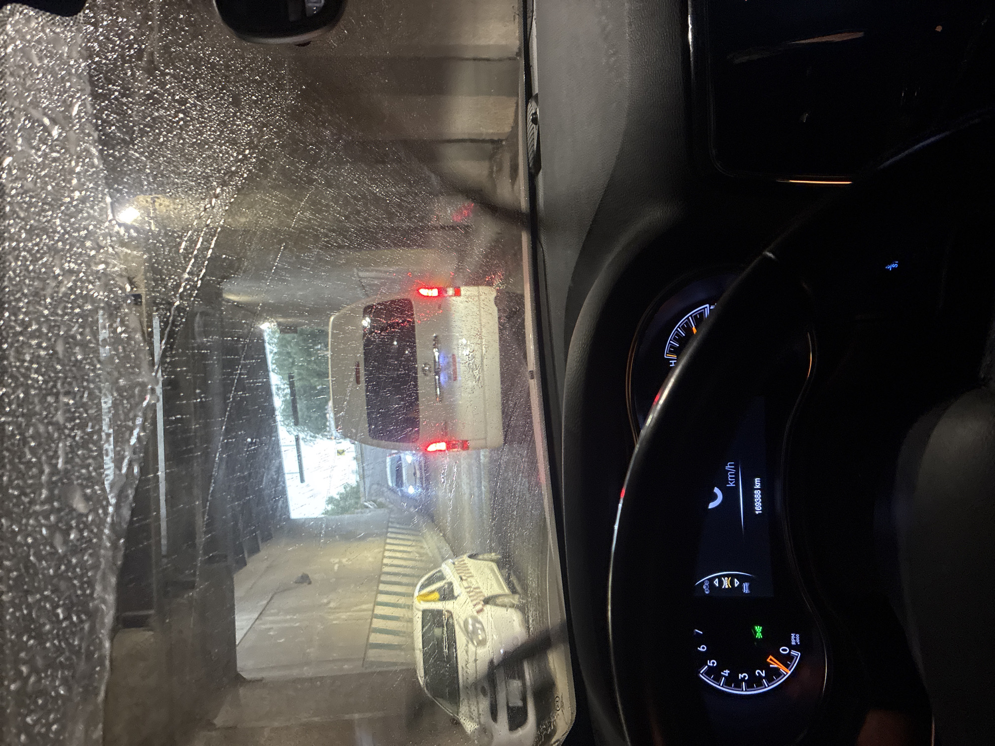 The image shows a view from inside a car going through a car wash in Cuautitlán Izcalli, Mexico. The windshield is covered in water droplets. In front of the car, other vehicles are visible through the wet windshield, one appearing to be a white van with its tail lights illuminated and another appears to be a taxi. The vehicle's dashboard is partially visible, showing the instrument cluster indicating the car is stationary with 169388 km mileage.