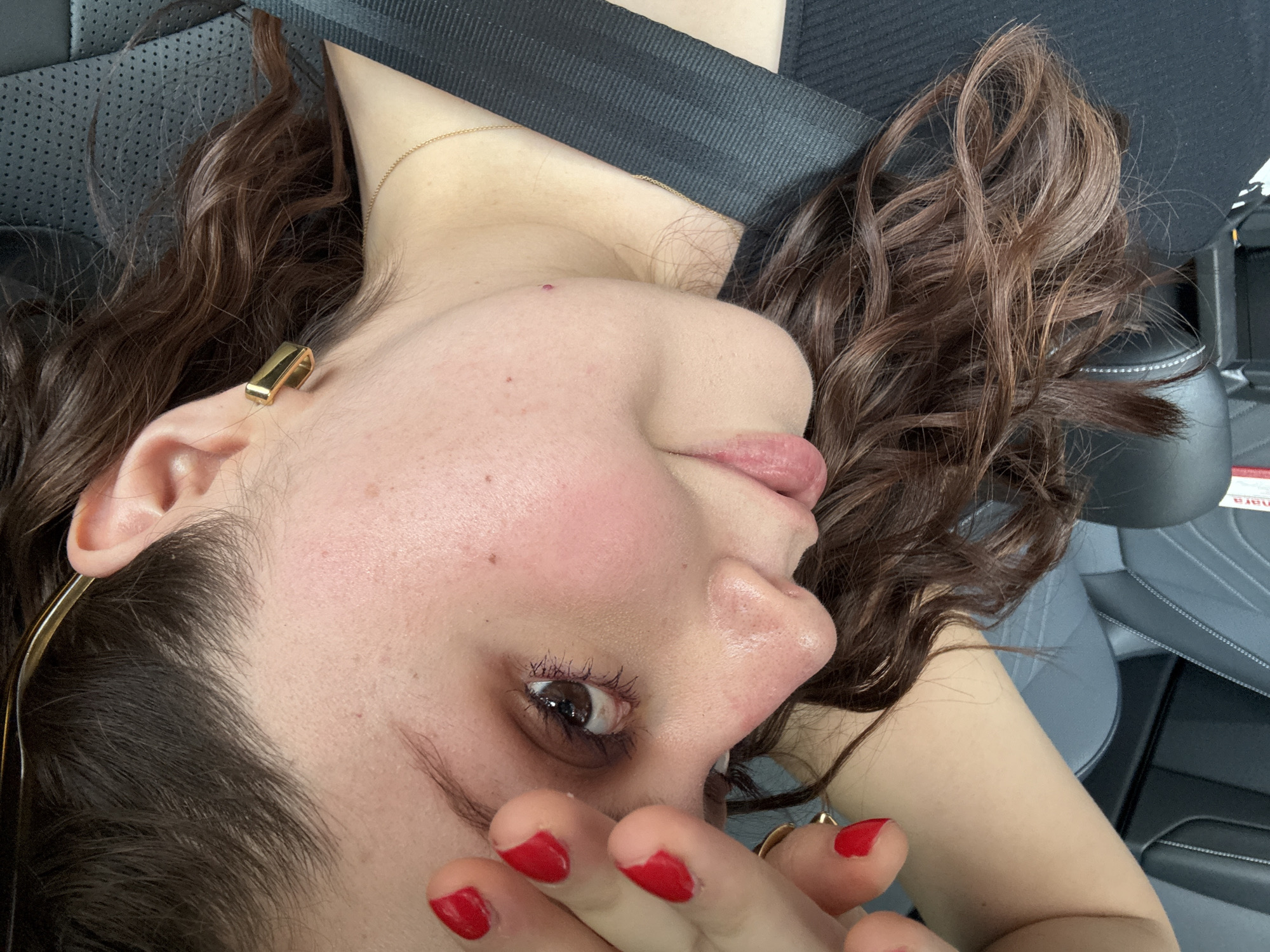 A woman is reclining in the front passenger seat of a car. She has curly brown hair, fair skin, and is wearing a black top and a seatbelt. She has red nail polish and is holding her hand up near her face. She wears a gold necklace. The interior of the car features black leather seats. The location is in Mexico City, Mexico, based on the text on the car window.
