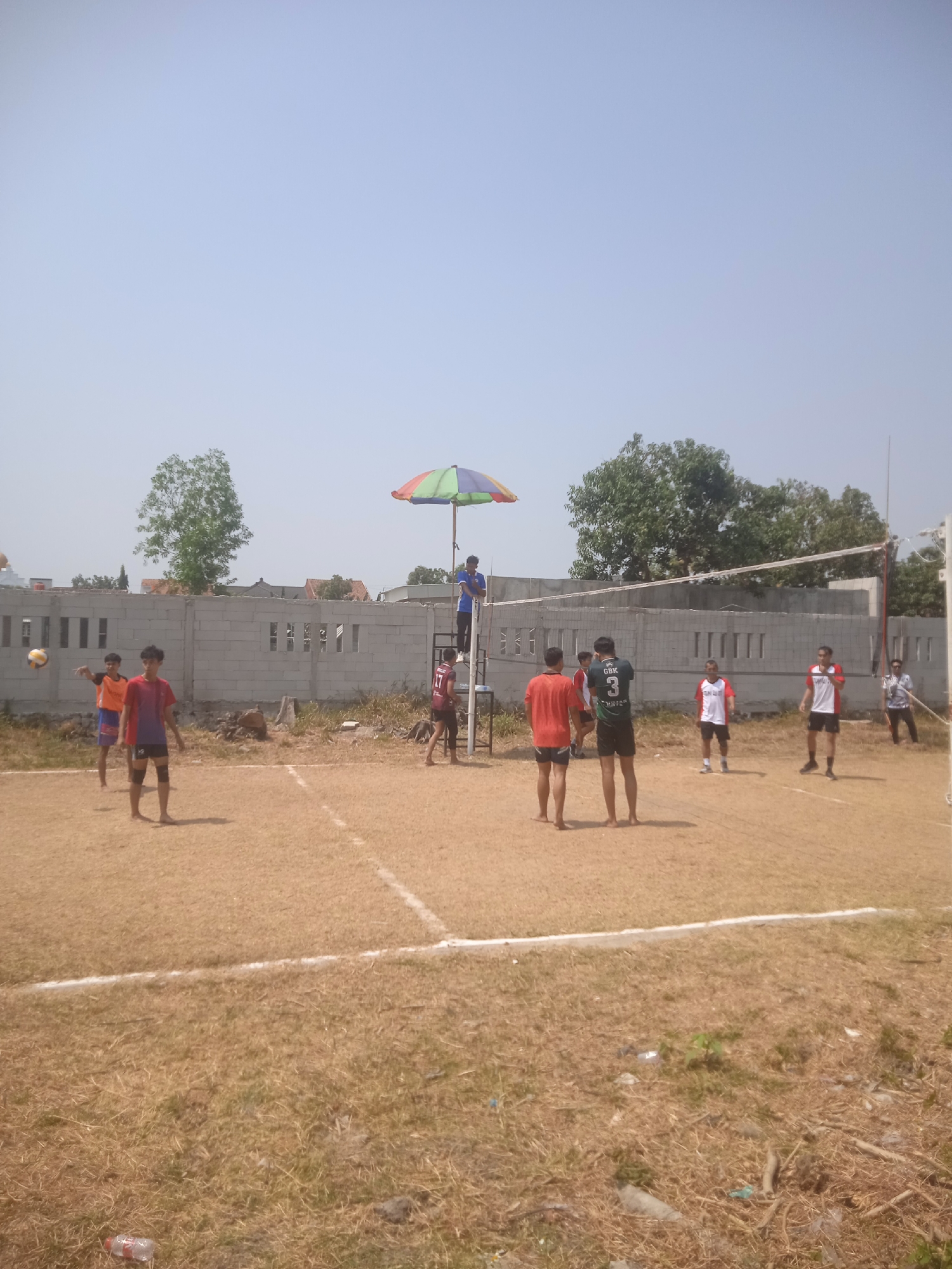 The image depicts an outdoor volleyball game in Klodran, Indonesia. Several young men are on a sand court, with a volleyball net stretched across it. One player is in the process of serving the ball. A referee sits in a high chair with an umbrella for shade. In the background, a low, cinder block wall separates the court from a residential area with houses and trees. The sky is clear and the weather appears to be sunny.