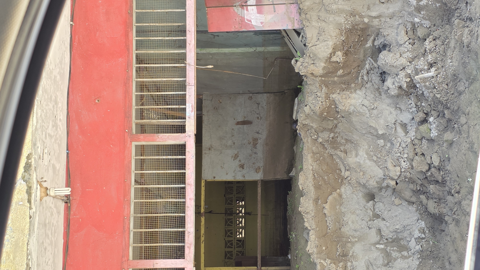 The image shows a building in Manyang Cut, Indonesia undergoing construction. A red facade is visible on the left with a barred window. Next to the building, a dirt excavation pit is present. The building's interior is dimly lit and appears unfinished. The ground adjacent to the building is disturbed with exposed soil and rocks.