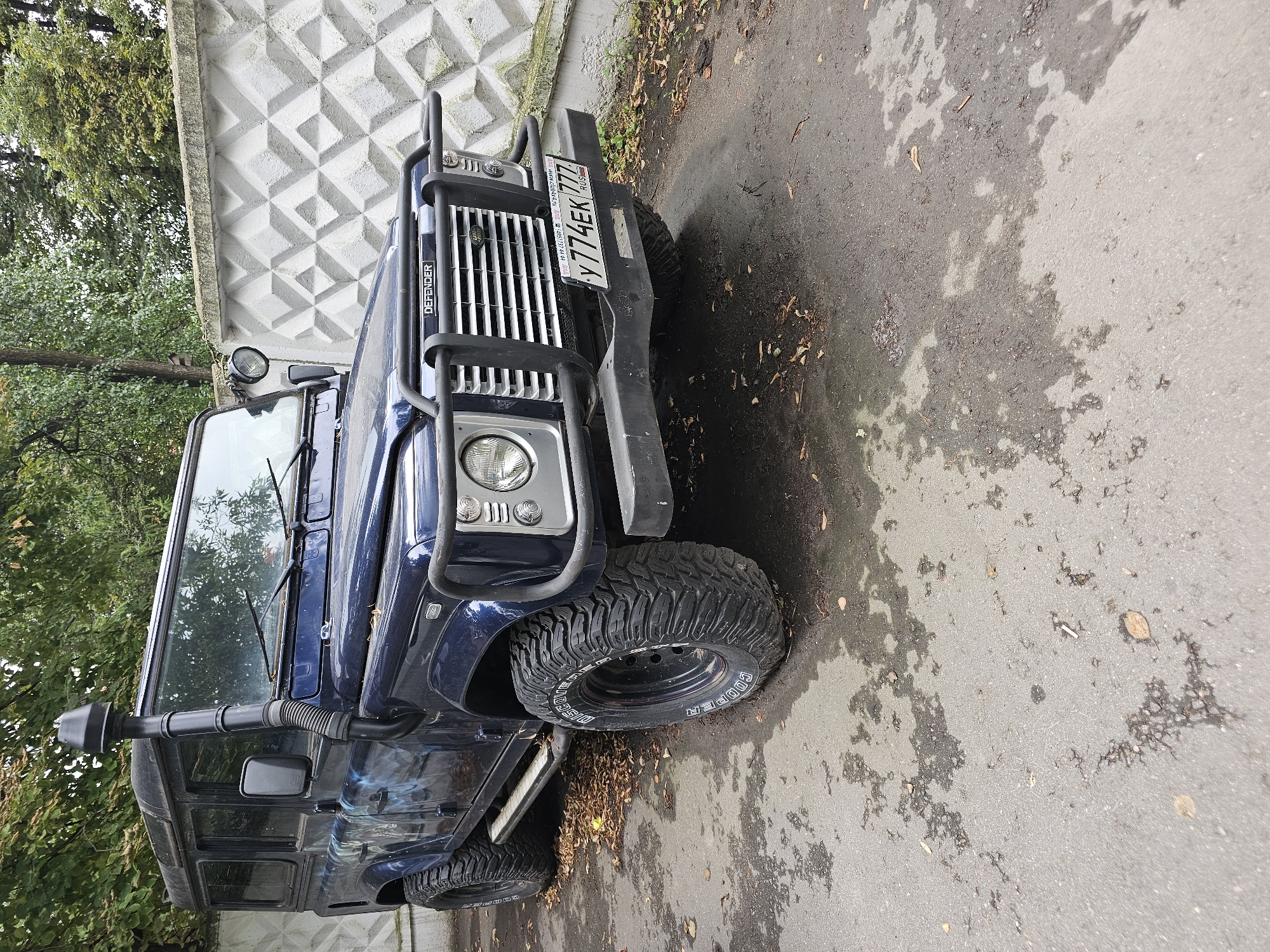 A dark blue Land Rover Defender is parked on a wet asphalt surface in front of a textured concrete wall, with trees and foliage in the background. The vehicle has a snorkel intake on the driver's side, large off-road tires, and a bull bar on the front. A Russian license plate reading "Y774EK 777" is visible on the front bumper. Fallen leaves are scattered on the ground around the vehicle. The scene appears to be a street or parking area in Moscow, Russia, given the license plate format and the appearance of the wall and surrounding vegetation.