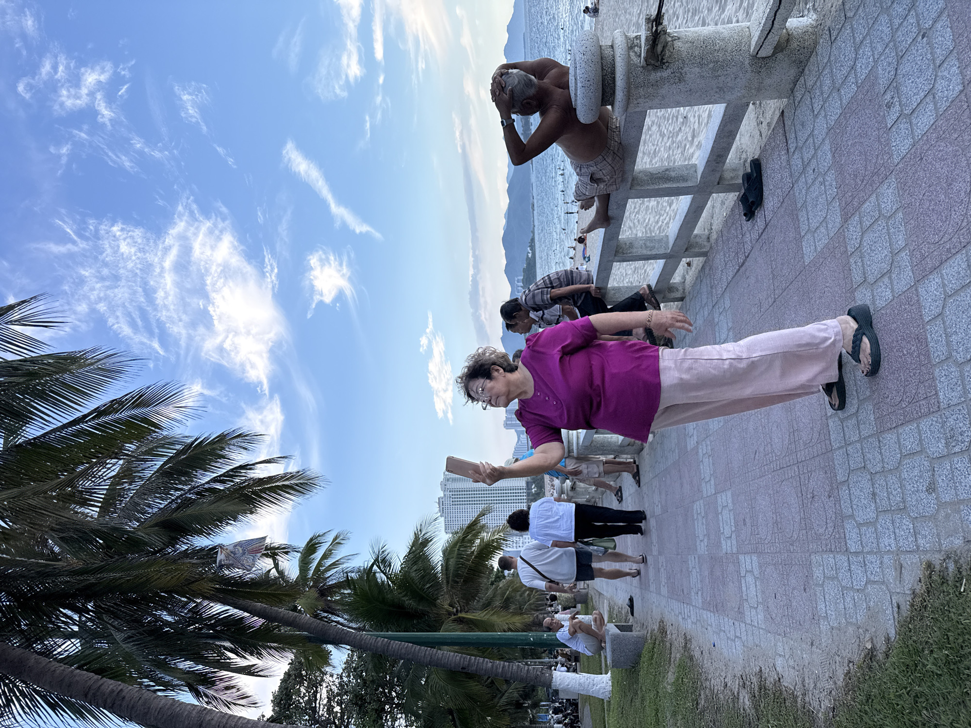 In this low-angle, wide shot, a group of people are seen on a paved walkway next to a beach. The sky above is a vibrant blue, dotted with wispy white clouds, suggesting a clear day. Palm trees with their fronds in the foreground frame the scene, hinting at a tropical location. In the background, the calm ocean stretches to the horizon, meeting a hazy mountainous coastline. Buildings can be seen in the distance, suggesting a developed area.
The primary focus is on an older woman in the foreground, wearing glasses and a bright magenta top with loose-fitting light pink pants. She is holding a smartphone and appears to be taking a photo or video, looking towards the right side of the frame with a neutral expression. To her right, several other individuals are also present on the walkway. Further back, a man is seated on a concrete structure near the beach, with his hands clasped behind his head. He is wearing plaid shorts and is shirtless. Other people are visible on the beach and further along the walkway, some standing and others seated, engaged in various activities. Two pairs of dark flip-flops are placed on the tiled walkway near the concrete structure. The time of day appears to be late afternoon, given the angle of the sun and the quality of the light. The overall atmosphere seems relaxed and casual, consistent with a day at the beach.