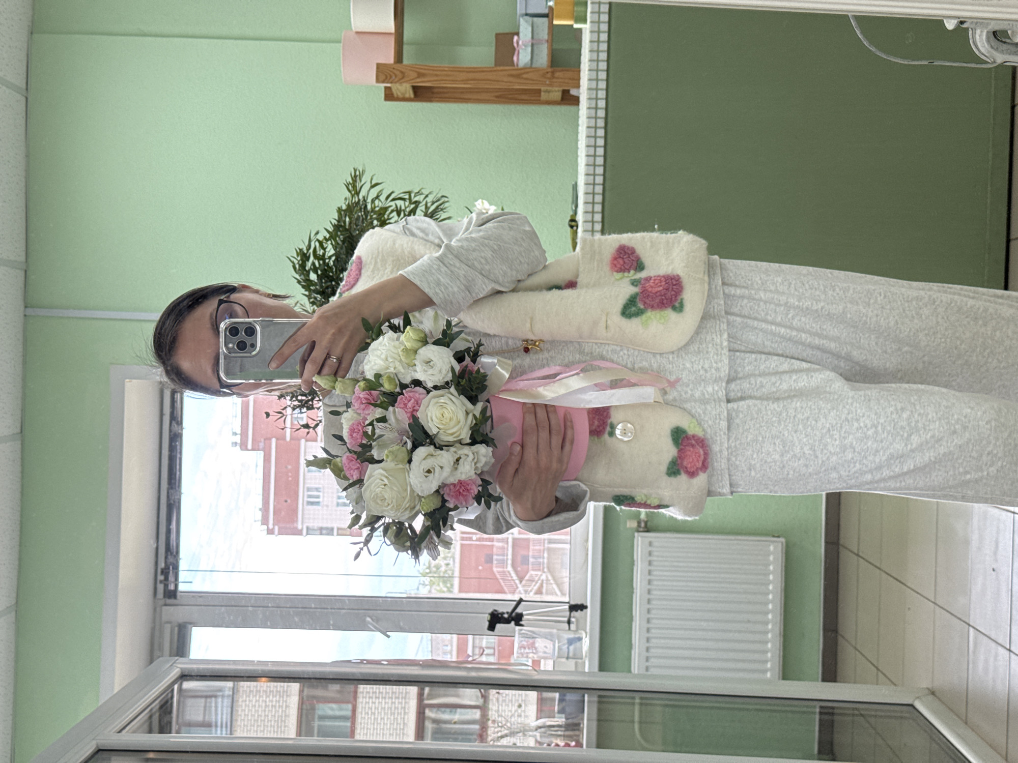 A woman, seen through a mirror reflection, captures a selfie while holding a vibrant bouquet and a pair of unique felt slippers. She wears dark-rimmed glasses and a light-colored long-sleeved top, her right hand holding the smartphone to take the picture. A ring is visible on her finger. Cradled in her left arm is a bouquet featuring an assortment of white roses and pink carnations or similar flowers, adorned with pink ribbons. Resting on her front are a pair of cream-colored felt slippers, meticulously embroidered with striking pink and green floral designs, resembling roses.
The indoor setting includes a room with light green walls. Above the woman's reflected head, a wooden shelf holds rolls of paper or small wrapped packages. A large window, partially visible behind her, looks out onto an urban landscape with a building constructed of reddish-brown brick, featuring numerous white-framed windows. A white radiator is mounted on the wall below the window.
The activity suggests a personal moment, possibly celebrating a gift, showcasing handmade items, or documenting a special occasion during the daytime. The handcrafted appearance of the felt slippers is a notable detail. The scene is situated in Izhevsk, Russia. No discernible text is visible.