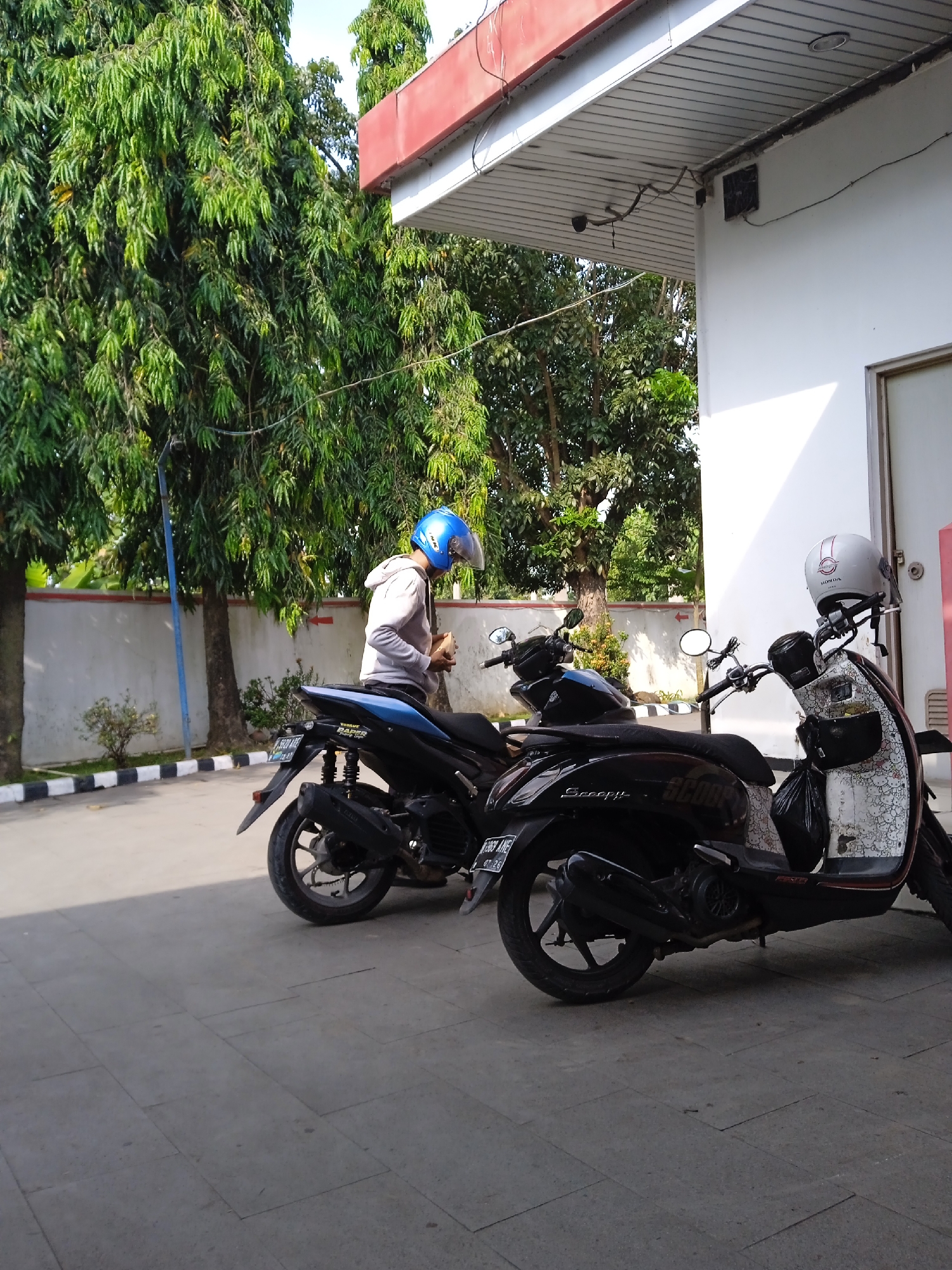 The image shows a scene in Katonsari, Indonesia. There are three parked motorcycles. A person with a blue helmet and a light grey hoodie stands next to the first motorcycle. The second motorcycle is black with "Scoopy" written on it. The third scooter is decorated with a Hello Kitty pattern. They are parked near a white building with a red awning. Trees and bushes are visible in the background.