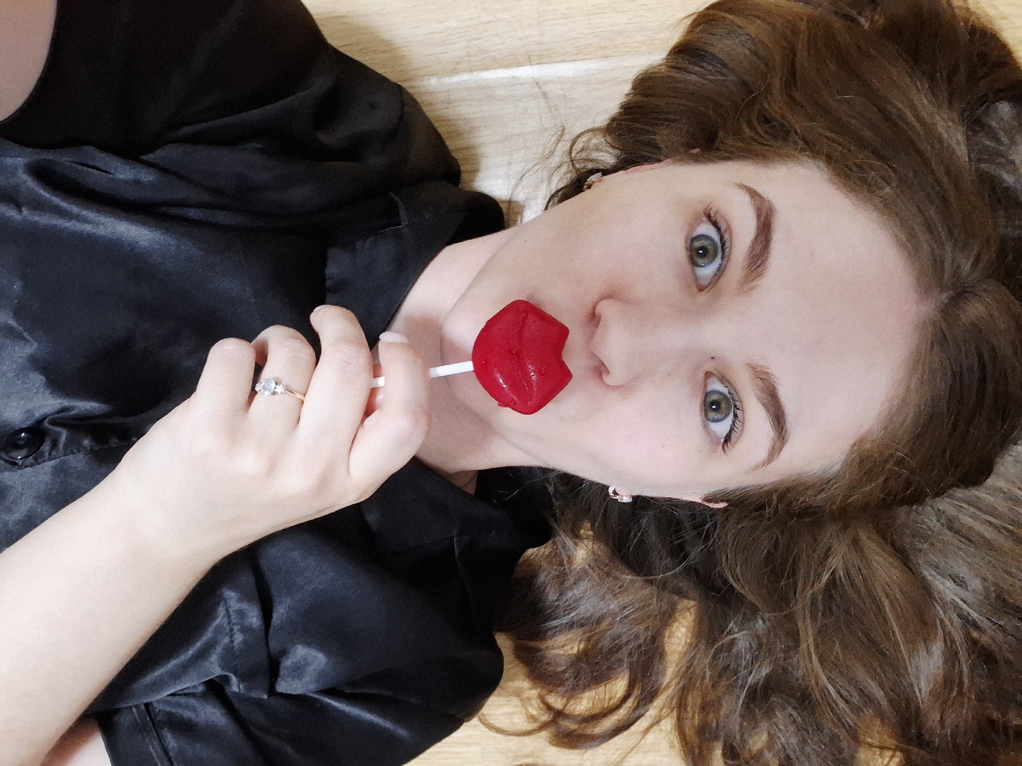 A young woman is lying on a wooden floor in an indoor location. She has light skin, brown hair, and green eyes. She is wearing a shiny black blouse. She holds a red, lip-shaped lollipop in her left hand and places it near her mouth. She wears a ring on her left ring finger. A small earring is visible in her right ear. The image provides no information regarding the specific location in Chaikovsky, Russia.