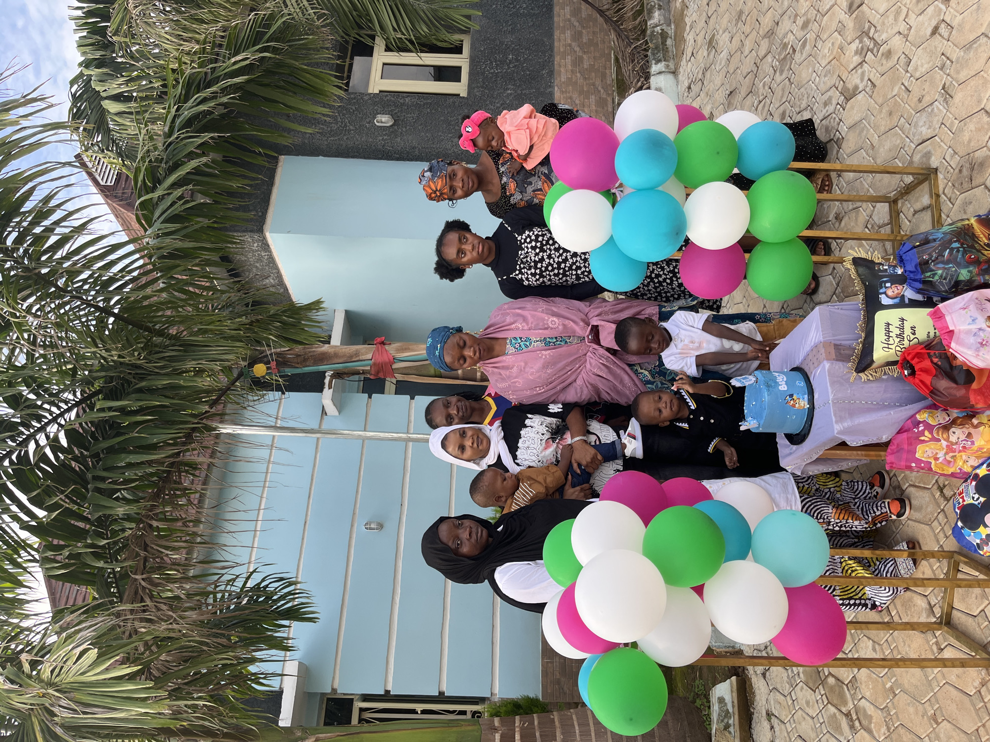 The scene is set outdoors in the city of Jos, Nigeria, featuring a group of ten individuals, predominantly women and children, gathered in front of a residential building. The ground is paved with interlocking hexagonal stones.
The group consists of six adult females and four children. On the left, a woman in a black hijab and white top holds an infant boy in a brown striped shirt. Next to her, a woman in a white hijab and patterned black top holds another infant boy in a brown shirt. To her right, a young girl in a black long-sleeved top and patterned dress stands behind. Further right, a woman in a multi-colored headwrap and patterned dress holds an infant girl in a pink outfit and a pink bow headband. Another woman is partially visible behind them in a black and yellow top. On the far left, a woman wears a blue patterned headscarf and a pink shawl over a patterned dress.
In the foreground, two young boys stand near a small table. The boy on the left wears a white polo shirt, and the boy on the right wears a dark polo shirt with gold trim. On the table, draped with white fabric, rests a light blue two-tier birthday cake adorned with cartoon characters and "Happy Birthday" text. Around the table, several colorful gift bags featuring cartoon characters (Disney Princess, Mickey Mouse, Spiderman) and a decorative pillow with "Happy Birthday Son" text are placed on the ground.
Two tall golden stands holding clusters of balloons in green, white, pink, and light blue colors frame the central area.
The background features a two-story residential building with light blue and dark grey textured walls and a brown roof. White trim is visible on the light blue section. Large palm trees with some dried fronds frame the upper part of the image and the building. The sky is partially visible through the foliage.