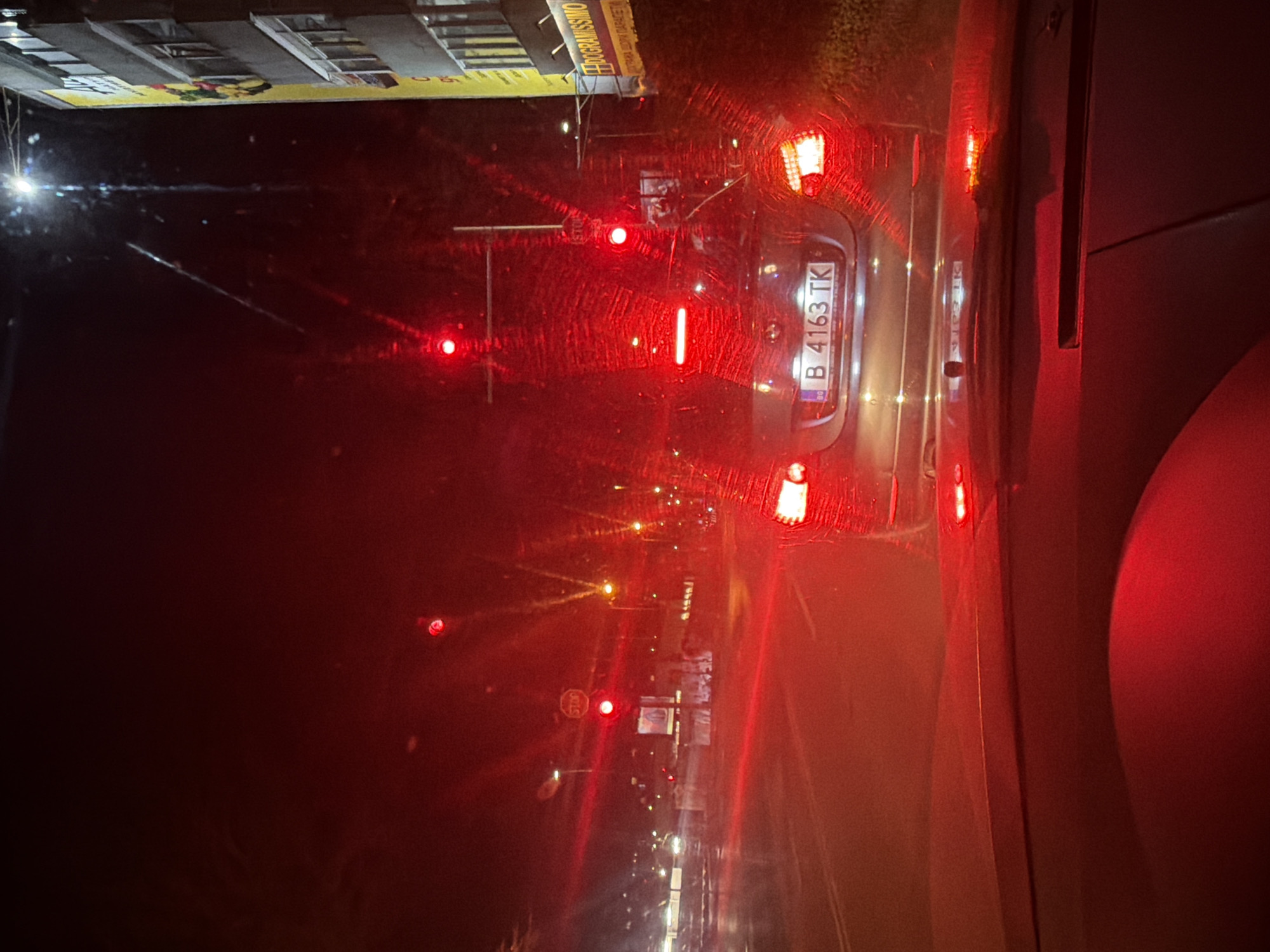 The image shows a nighttime scene from inside a car, looking out through the front windshield, which is wet from rain. The car is stopped at a traffic light. In front, there is another car with the license plate "B 4163 TK," which is a Bulgarian license plate. The red taillights and brake lights of the car in front are illuminating the scene. Streetlights and other car lights are visible in the distance, causing reflections on the wet windshield. On the left side of the image, there appears to be a sign or advertisement. The overall impression is of a rainy night in a city, possibly Varna, Bulgaria, based on the license plate. The image captures a common urban driving situation.