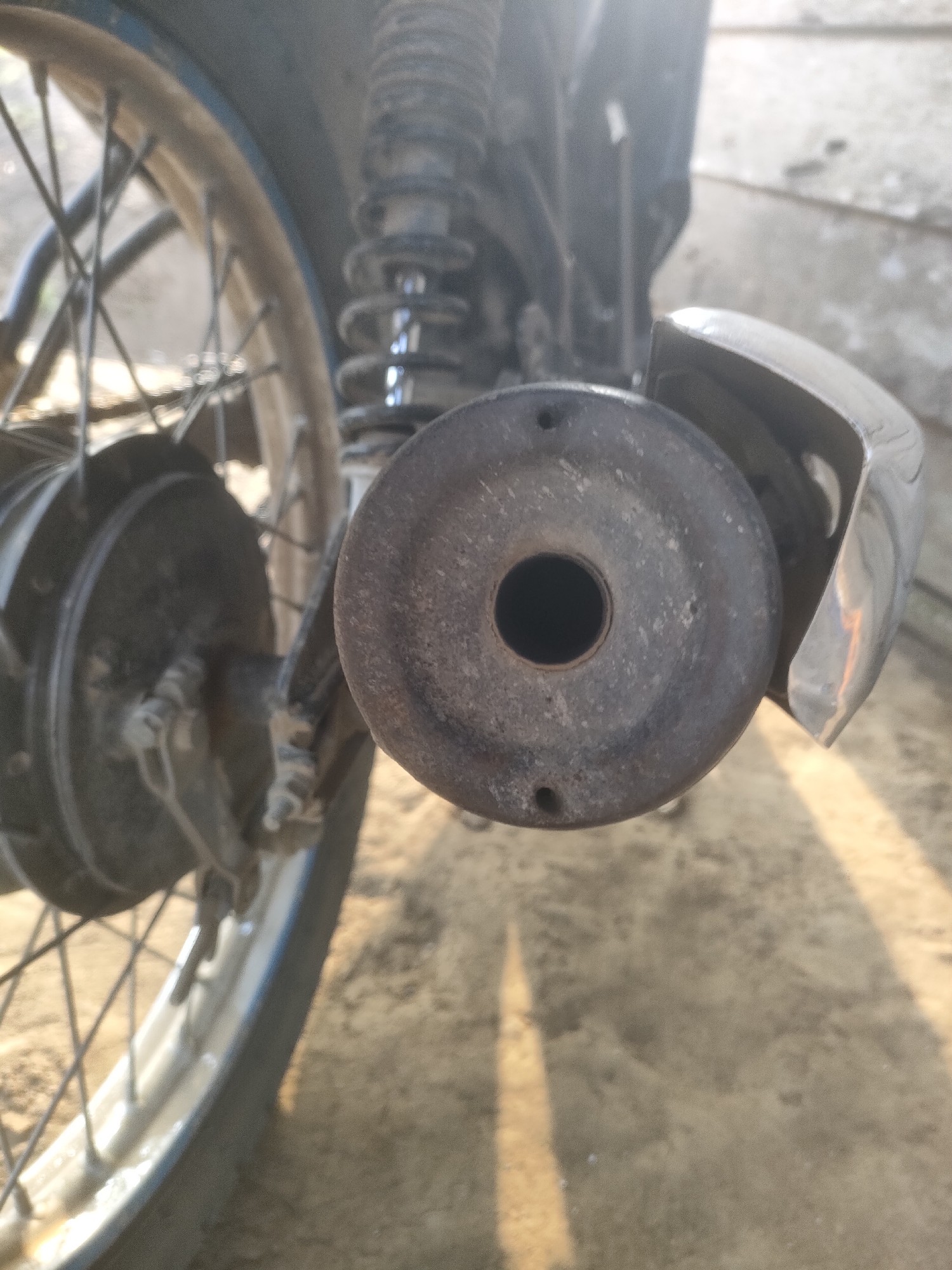 The image displays a close-up view of the rear of a motorcycle, focusing on the exhaust pipe and wheel. The exhaust pipe is circular with a dark, weathered appearance, showing visible wear and some small holes. Behind it, a chrome-plated fender is partially visible. To the left, a portion of the rear wheel with spokes and a tire is in focus. The motorcycle is positioned outdoors, with a sandy or dusty ground surface in the foreground, upon which strong shadows are cast, suggesting sunlight. The background is blurred and appears to be an outdoor environment, possibly a street or yard. There are no people or discernible activities captured in this image. The specific location of Padang Dalom, Indonesia, cannot be confirmed or denied solely from the visual information presented in the image.