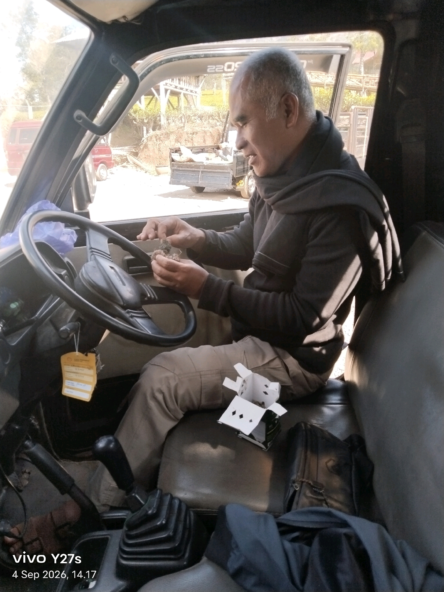 In the driver's seat of a truck in Sigedang, Indonesia, a man with short gray hair wears a gray scarf and a dark sweater. He holds a small, brown object in his hands and appears to be examining it. Next to him is an open white paper box with diamond-shaped cutouts. The interior of the truck is visible, including a steering wheel, gear shifter, and dark leather seats. Outside the truck, a red van and a trailer loaded with goods are partially visible. The background shows trees and a hillside. The timestamp on the image indicates it was taken on September 4, 2026, at 2:17 PM.