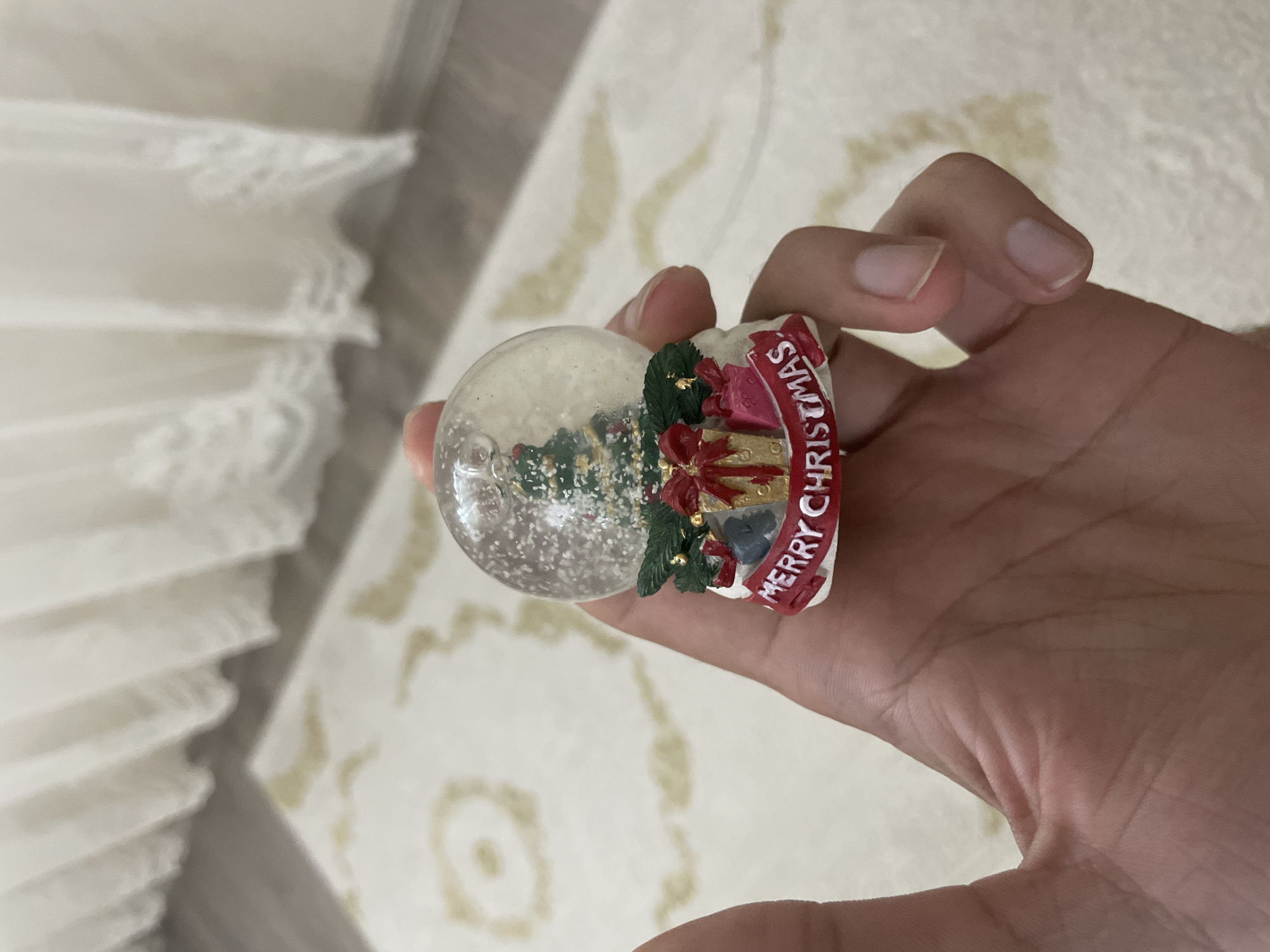 A close-up image shows a person's light-skinned hand holding a small, festive snow globe indoors in Ihtiman, Bulgaria. The hand is positioned centrally, with fingers curled around the decorative base of the snow globe.
The snow globe features a clear glass dome containing a miniature green Christmas tree amidst scattered white "snow" particles. Its base is intricately decorated with holiday elements: green foliage, small wrapped gift boxes (one red, one golden), and a red banner. The text "MERRY CHRISTMAS" is clearly visible in white lettering on this red banner, wrapping around the base.
In the background, to the left, a white, pleated curtain, possibly sheer, suggests a window and a source of natural light. The remaining background consists of a light-colored rug or carpet adorned with a faint, light brown, swirling pattern. The overall lighting is bright, indicative of daytime. The image focuses on the detail of the holiday ornament and the hand holding it.