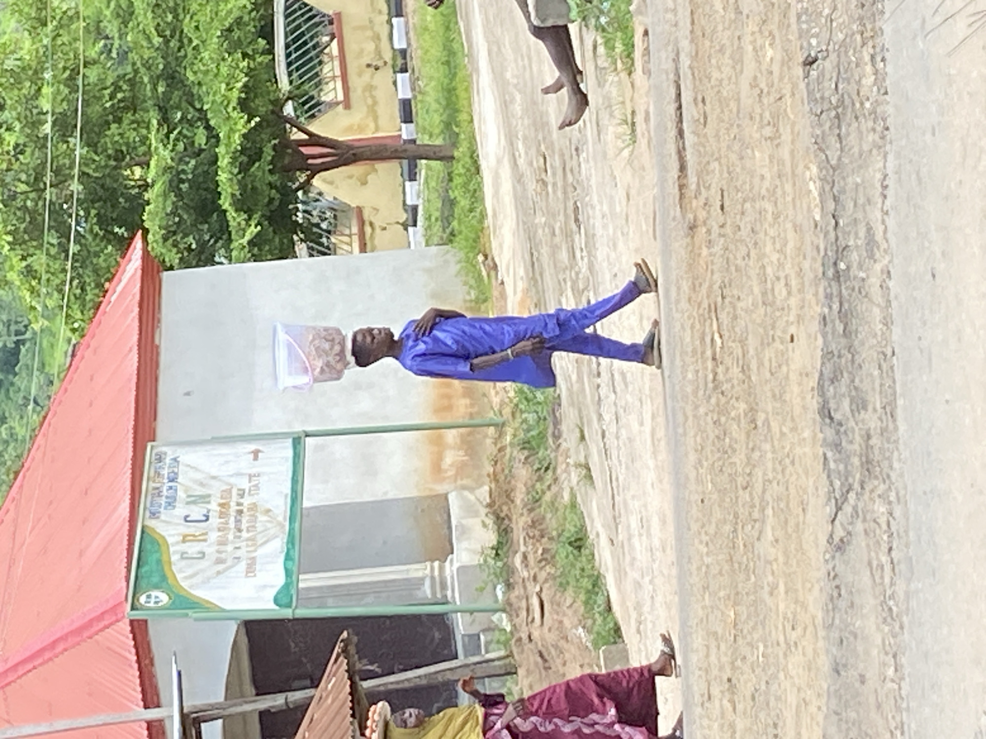 In Mararaba, Nigeria, a young person walks down a dirt road, carrying a bucket of goods on their head. The person is wearing a blue traditional outfit. To the left, a woman in a burgundy dress and yellow headscarf also walks down the road, carrying a basket of eggs on her head. In the background, a building with a red roof and a sign that reads "CRCN CHURCH MEGIDA" stands. The weather appears to be overcast.