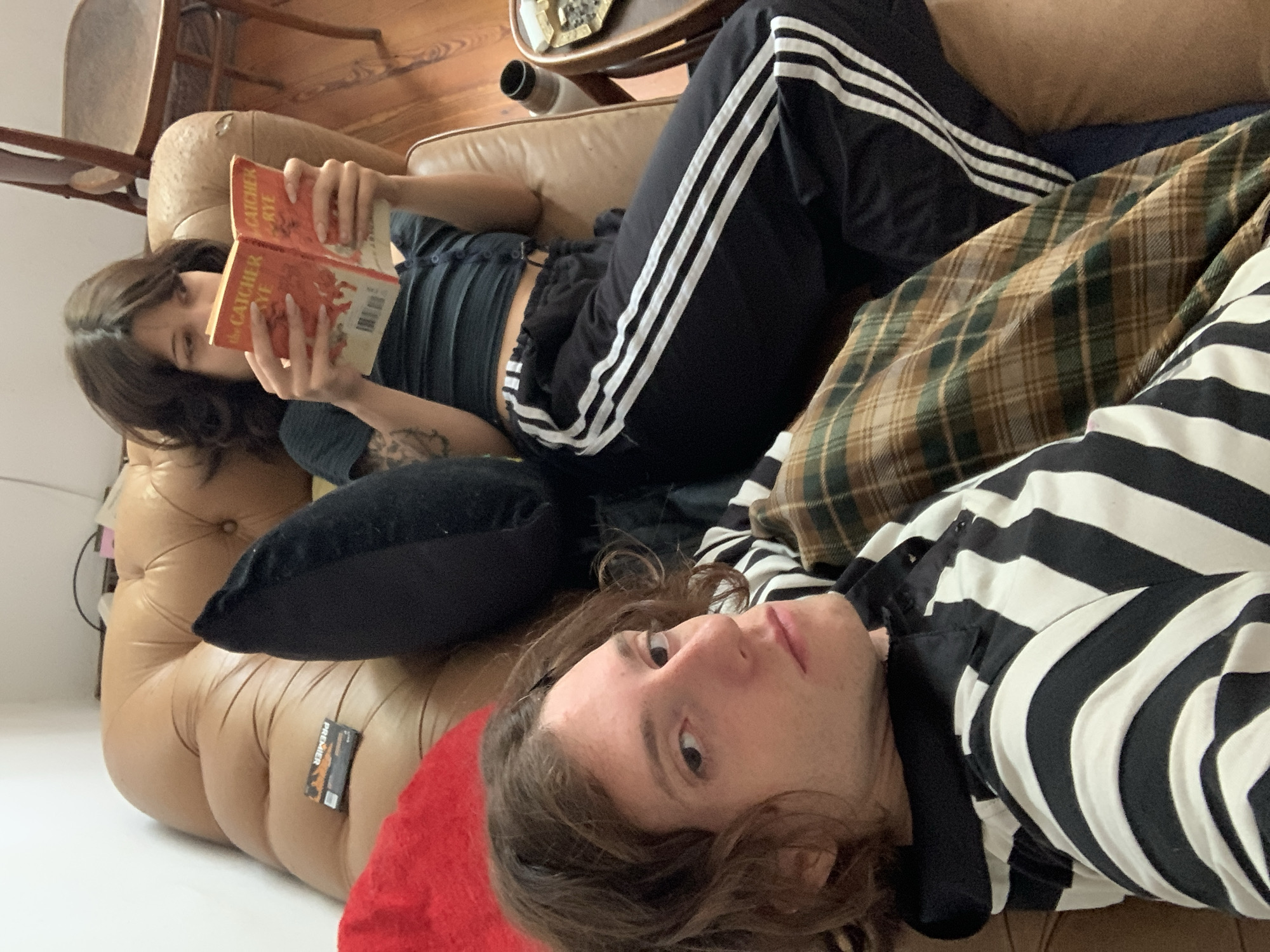 A young man and woman are lying on a tan leather sofa. The woman, positioned further away from the camera, is reading a book with a red cover, possibly titled "The Catcher in the Rye." She has dark hair and is wearing a dark top with visible straps and black pants with three white stripes down the side. The man, closer to the camera, is wearing a black and white striped shirt. A black pillow is behind the woman, and a brown and green plaid blanket lies near the man. A red cushion is visible under his head. A small black box with orange print is positioned on the leather sofa. A wooden chair is partially visible in the background on the left side. The scene appears to be indoors, possibly a home. The location is not evident to be in the city of Buenos Aires, Argentina. There are no visual indicators to confirm.