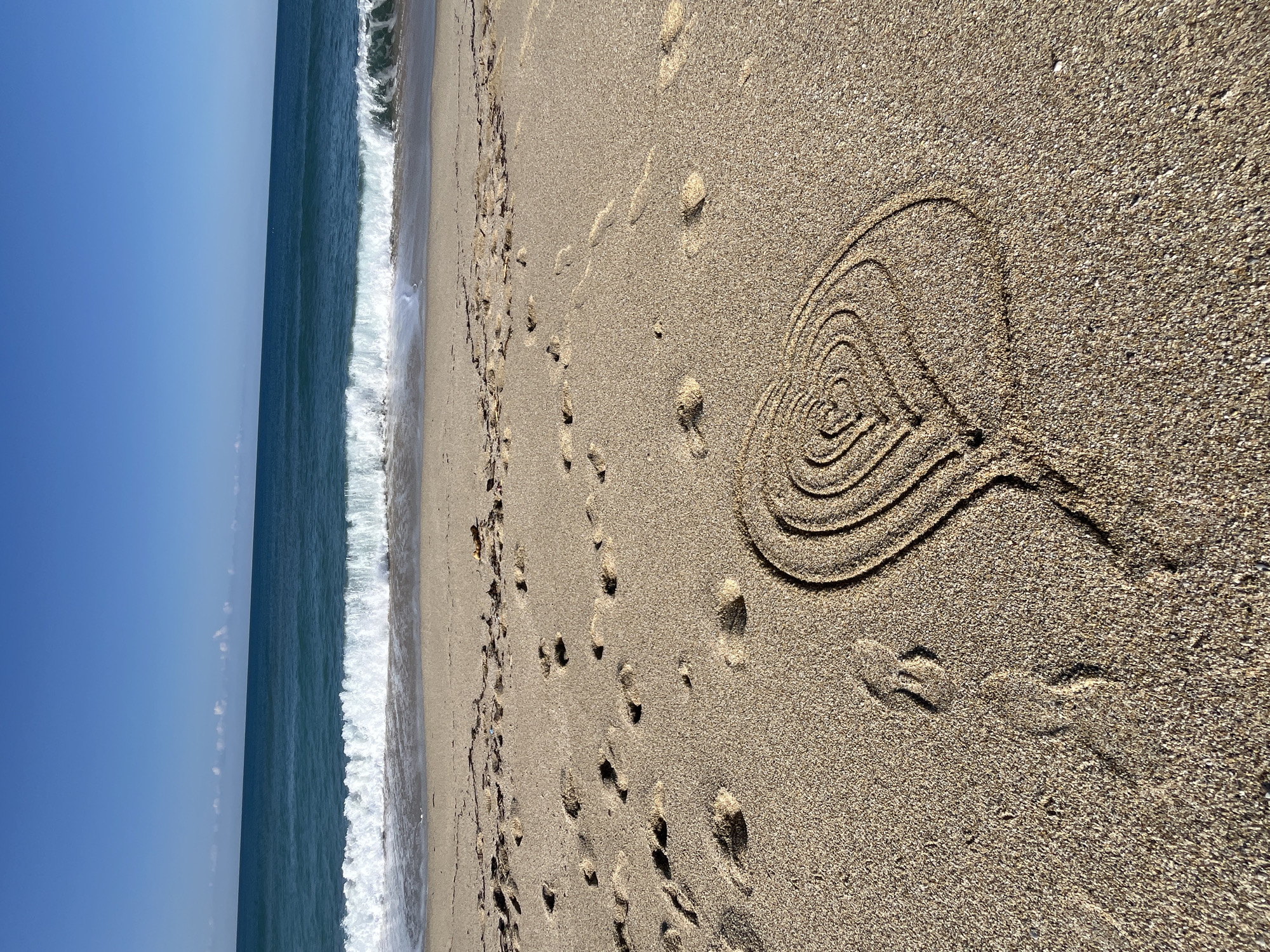 This is an outdoor shot of a sandy beach in Alanya, Turkey, alongside the sea. The sky is clear and blue. The sea stretches along the left side of the image, with gentle waves reaching the shore. The majority of the shot is a close-up of the sand, revealing numerous footprints scattered across its surface. On the right side, a heart-shaped figure is drawn into the sand.