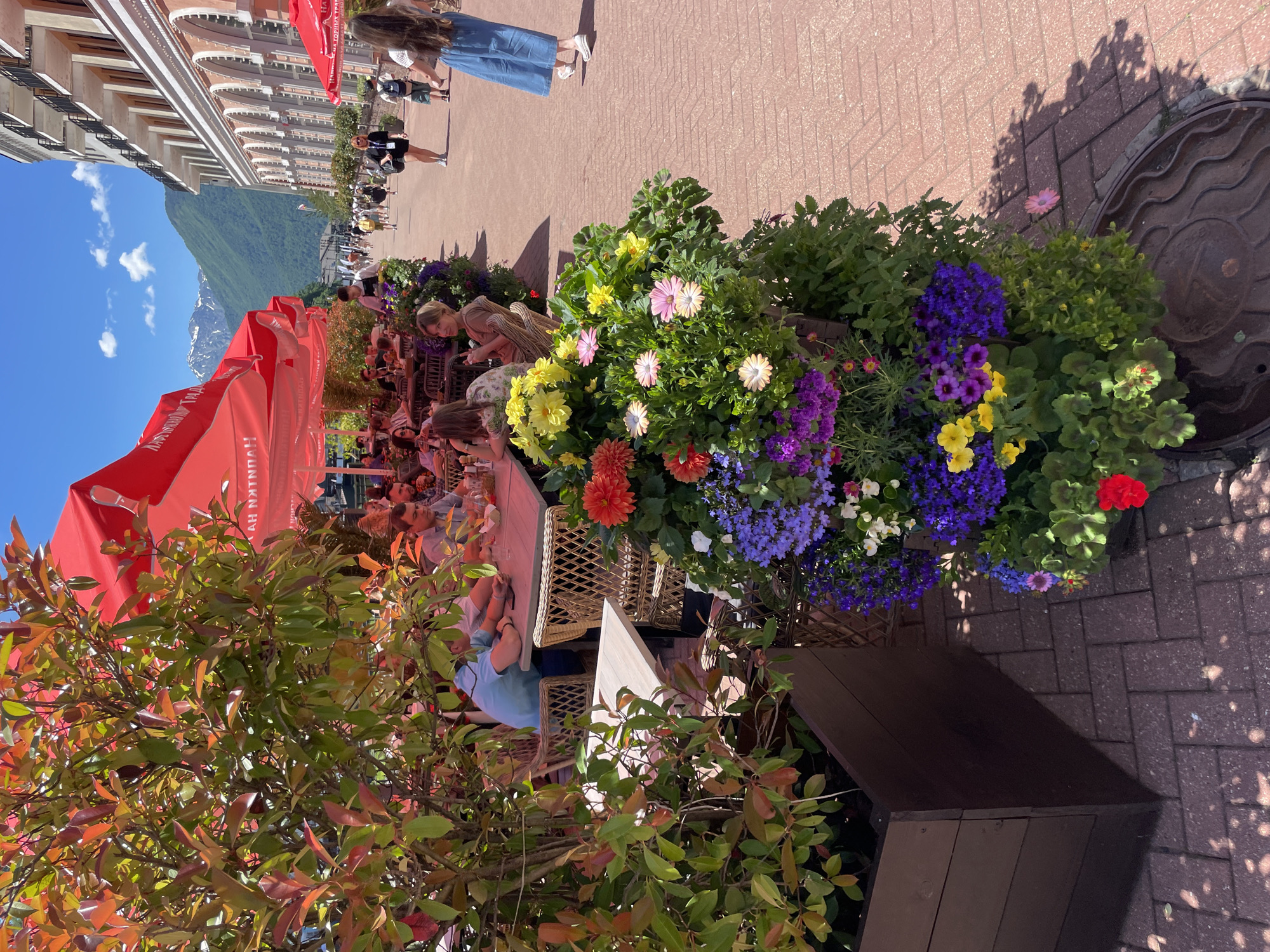 This image captures a street scene in Estosadok, Russia, likely during daylight hours. In the foreground, a large planter filled with vibrant flowers, including red, orange, yellow, pink, and purple blooms, is positioned on a brick-paved walkway. To the left, a wooden planter box and a tree with red and green leaves add to the lush greenery.
Further back, outdoor seating areas with tables and wicker chairs are set up under large red umbrellas. Several people are seated at these tables, appearing to dine or socialize. A few individuals are walking along the walkway, including a woman in a denim skirt and a young girl in a blue dress.
The background reveals a street lined with buildings, one of which has a classical facade with arched windows. In the distance, snow-capped mountains are visible against a bright blue sky with scattered clouds. The overall atmosphere suggests a pleasant day in a resort town.