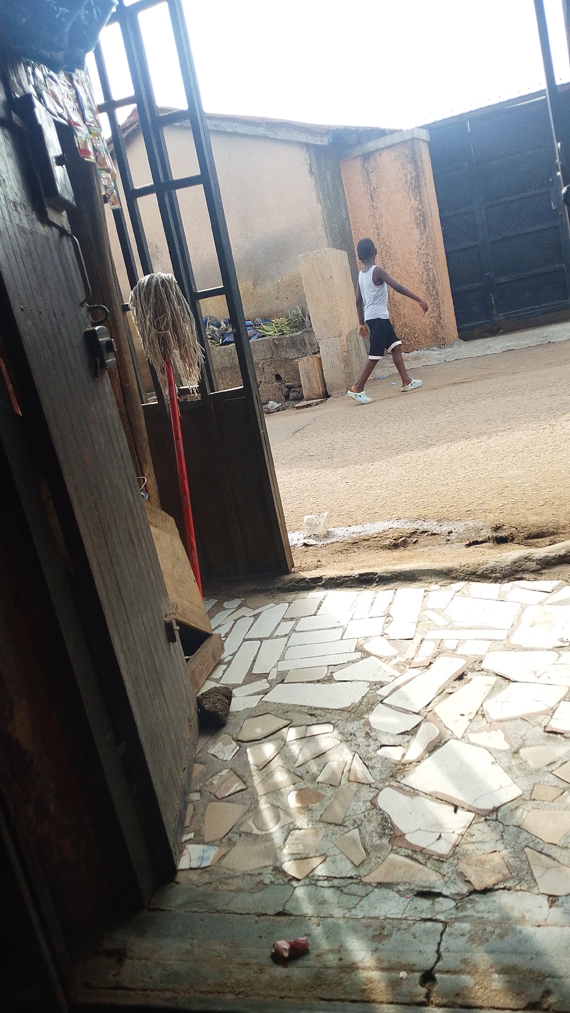 In an outdoor scene, a young boy walks away from the camera, heading towards a black gate. He is wearing a white tank top and black shorts, with white sneakers on his feet. The boy is on a paved surface that appears to be a street or walkway. To the left of the boy, there is a wooden structure, possibly a doorway or fence, and a mop with a red handle is leaning against it. In the background, there are buildings and a beige wall. Sunlight casts shadows on the ground. A piece of red meat is visible on the cracked pavement in the foreground.