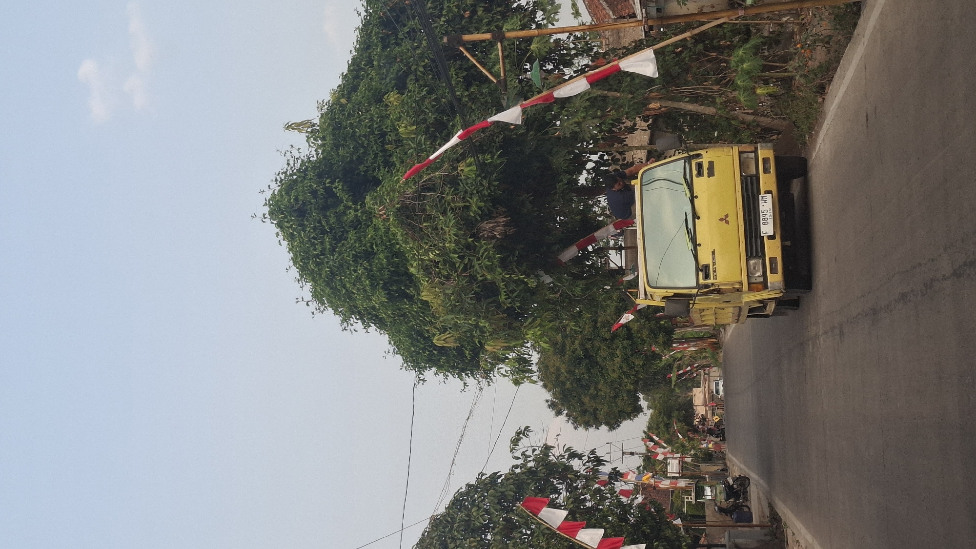 The image depicts a street scene in Cibinong Hilir, Indonesia, under a clear sky. A yellow Mitsubishi truck is prominently featured on the right side of the road, facing forward. A person, wearing a dark blue shirt, is visible on top of the truck's cabin. The road is paved and curves slightly to the left, leading into the distance. Along both sides of the street, trees with dense green foliage line the way, with red and white Indonesian flags adorning some of them and poles. A motorcycle is parked on the left side of the road near a building. In the background, more flags and houses are visible. The overall atmosphere suggests a festive or patriotic occasion.