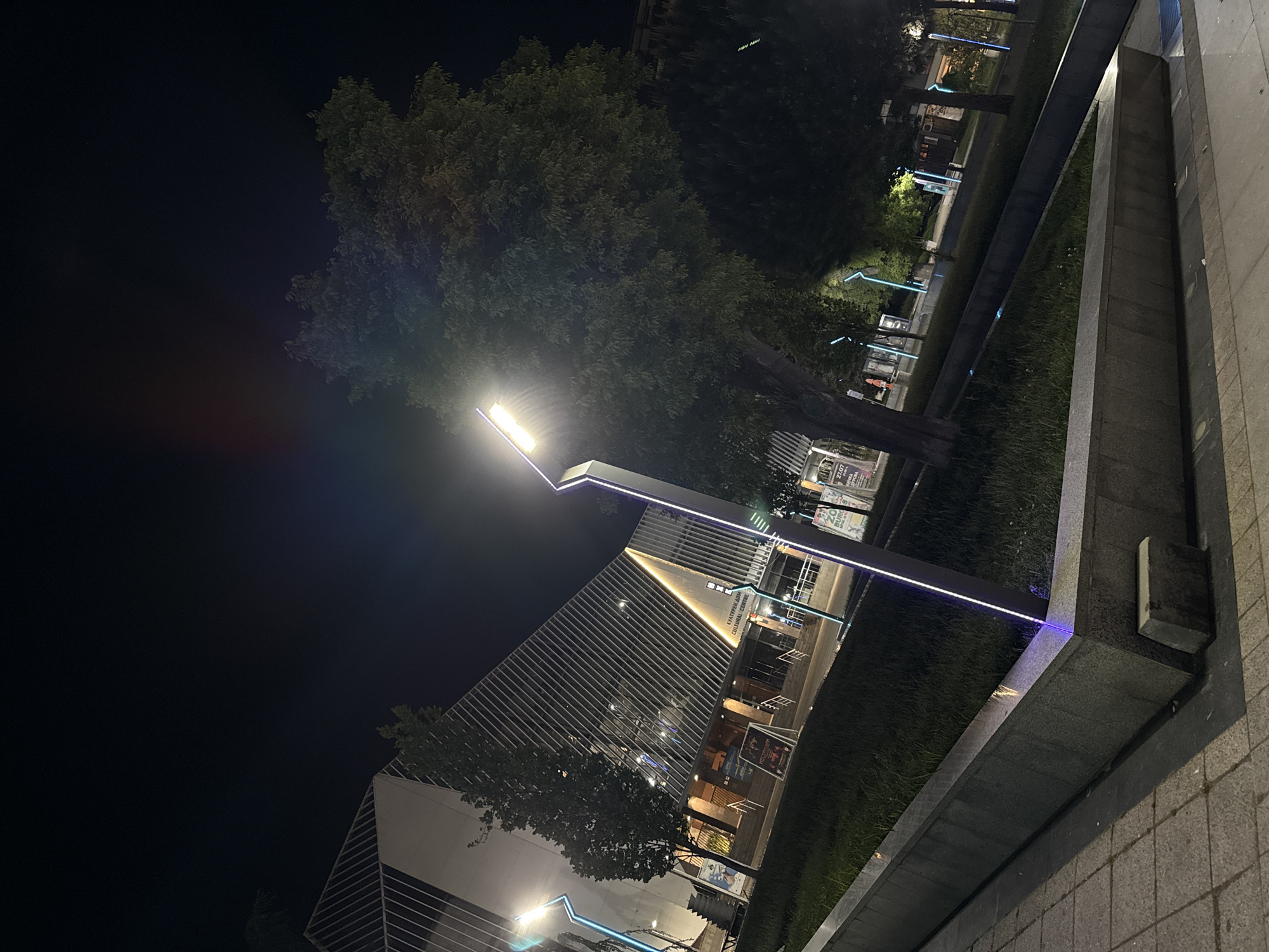 The image shows a night view of the Cultural Center in Burgas, Bulgaria. A modern building with a triangular facade featuring parallel lines is illuminated with internal lights. A streetlight with a minimalist design stands prominently in the foreground. A nearby tree frames the building. The area is landscaped with greenery. The sidewalk and a low concrete wall are visible in the foreground, leading towards the cultural center. Other illuminated structures, likely streetlights or signage, are visible further down the path. The night sky is dark, with some light glare around the streetlight.