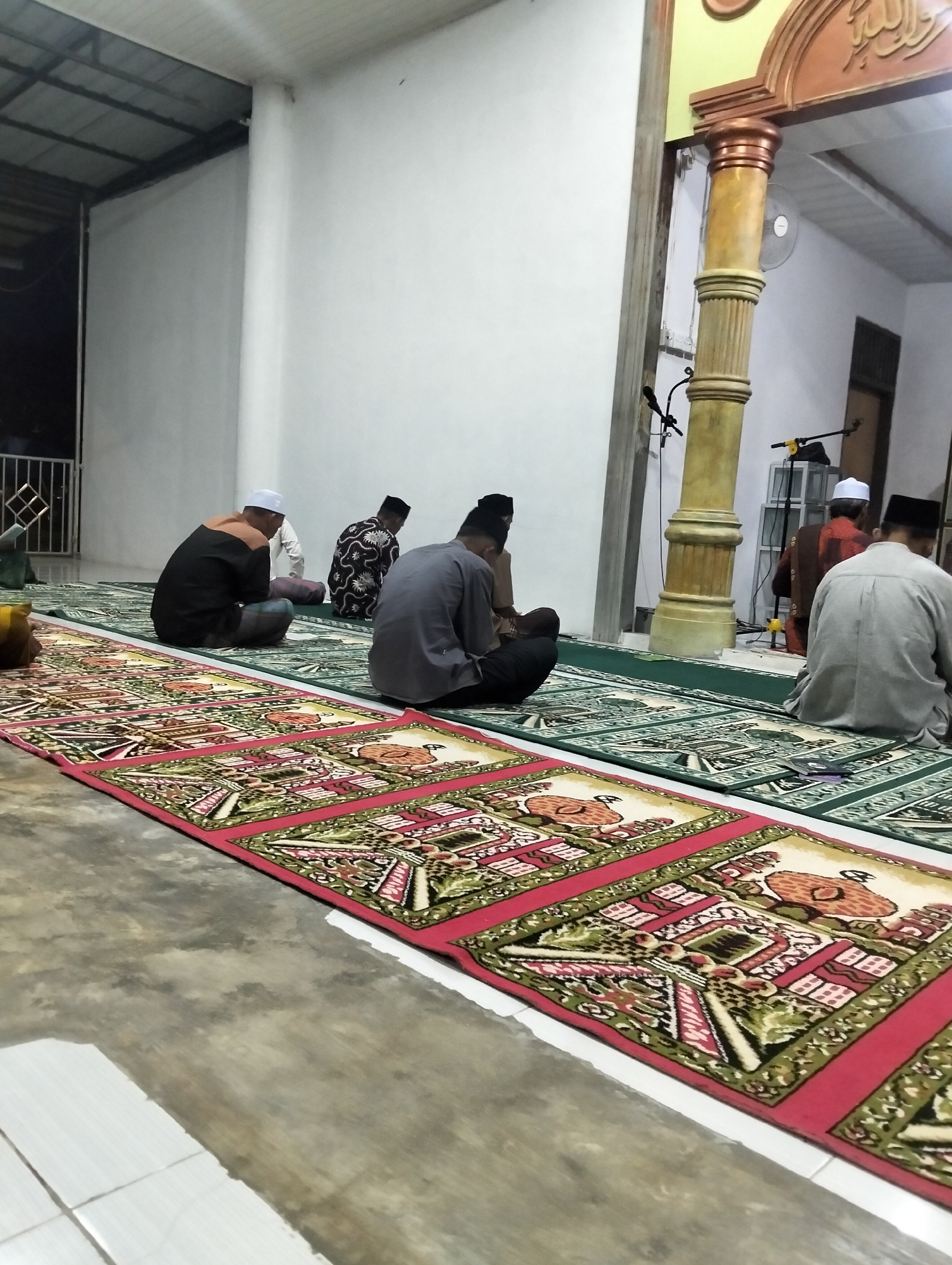 The image depicts a group of Muslim men engaged in prayer inside a mosque in Payagaboh, Indonesia. The men are seated on prayer rugs laid out on the floor. The setting appears to be an indoor space, possibly a hall or an extension of the mosque, with a concrete floor and white walls. Natural and artificial lighting illuminate the scene.
In the foreground, a man in a black shirt and a white cap is seen from behind, seated on a prayer rug with a red border and intricate floral and architectural patterns. To his left, another man in a patterned shirt and a black cap is also seated. Further back, a man in a grey shirt and black cap is in a prayer posture. In the middle ground, a man in a grey shirt and a white cap is seated. In the background, another man in a reddish-brown garment and a white cap is visible.
The men are all facing in the same direction, presumably towards the qibla, the direction of the Kaaba in Mecca, which is a standard practice in Islamic prayer. The atmosphere seems serene and devout.
Notable details include the ornate prayer rugs with their detailed designs, suggesting a cultural or religious significance. A large, ornate pillar with classical detailing stands to the right of the center, adorned with Arabic calligraphy at the top. A ceiling fan is visible behind the pillar. There are also microphones and lighting equipment in the background, indicating the potential for prayer services or religious gatherings. The time of day cannot be definitively determined, but the lighting suggests it could be late afternoon or evening, or an indoor space with artificial lighting. The location is identified as Payagaboh, Indonesia. No discernible emotions are overtly expressed, but the posture and context imply solemnity and devotion. No visible text is clearly legible in the image, apart from the Arabic calligraphy on the pillar.