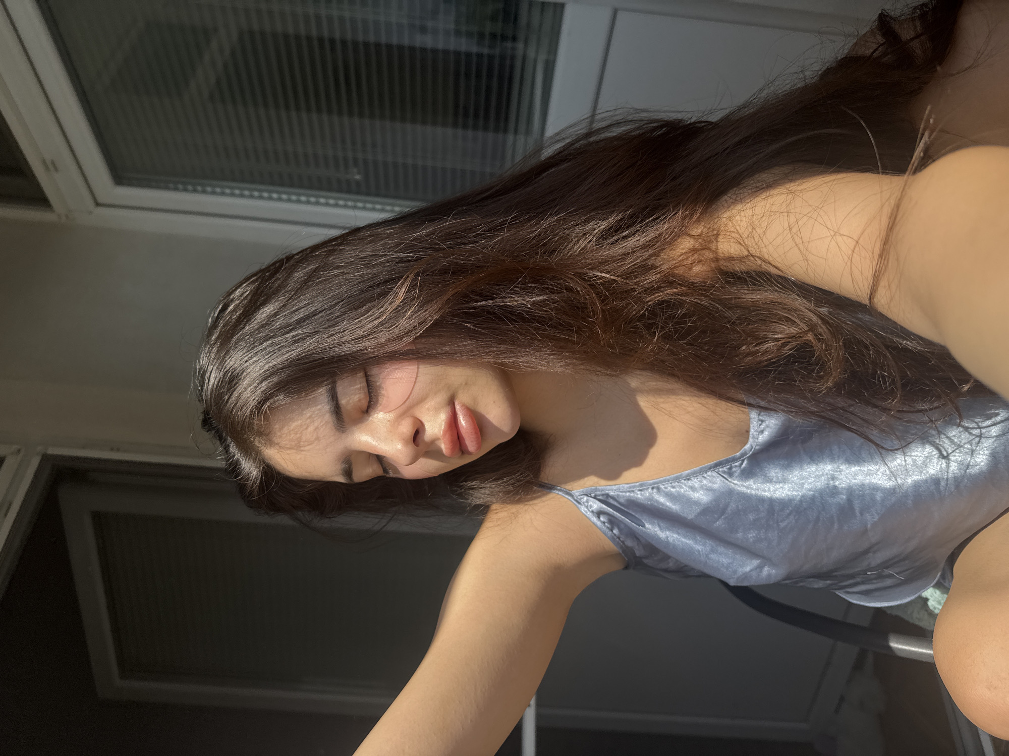 A close-up, portrait-oriented photograph captures a young woman illuminated by sunlight indoors in Plovdiv, Bulgaria. The woman, with long, flowing dark brown hair, has her eyes closed and a peaceful expression, suggesting relaxation or enjoyment of the warm light. Her head is tilted, and her skin glows under the direct sunlight, which casts soft shadows on her face and highlights her light blue or silver satin camisole. Her left arm is visible, extending diagonally downwards.
The background features parts of a window with horizontal blinds or louvers, indicating an indoor setting, possibly an apartment or a room with large windows or a balcony. The strong, low-angle sun suggests the time of day is either morning or late afternoon. The overall mood is calm and serene. There is no visible text in the image.