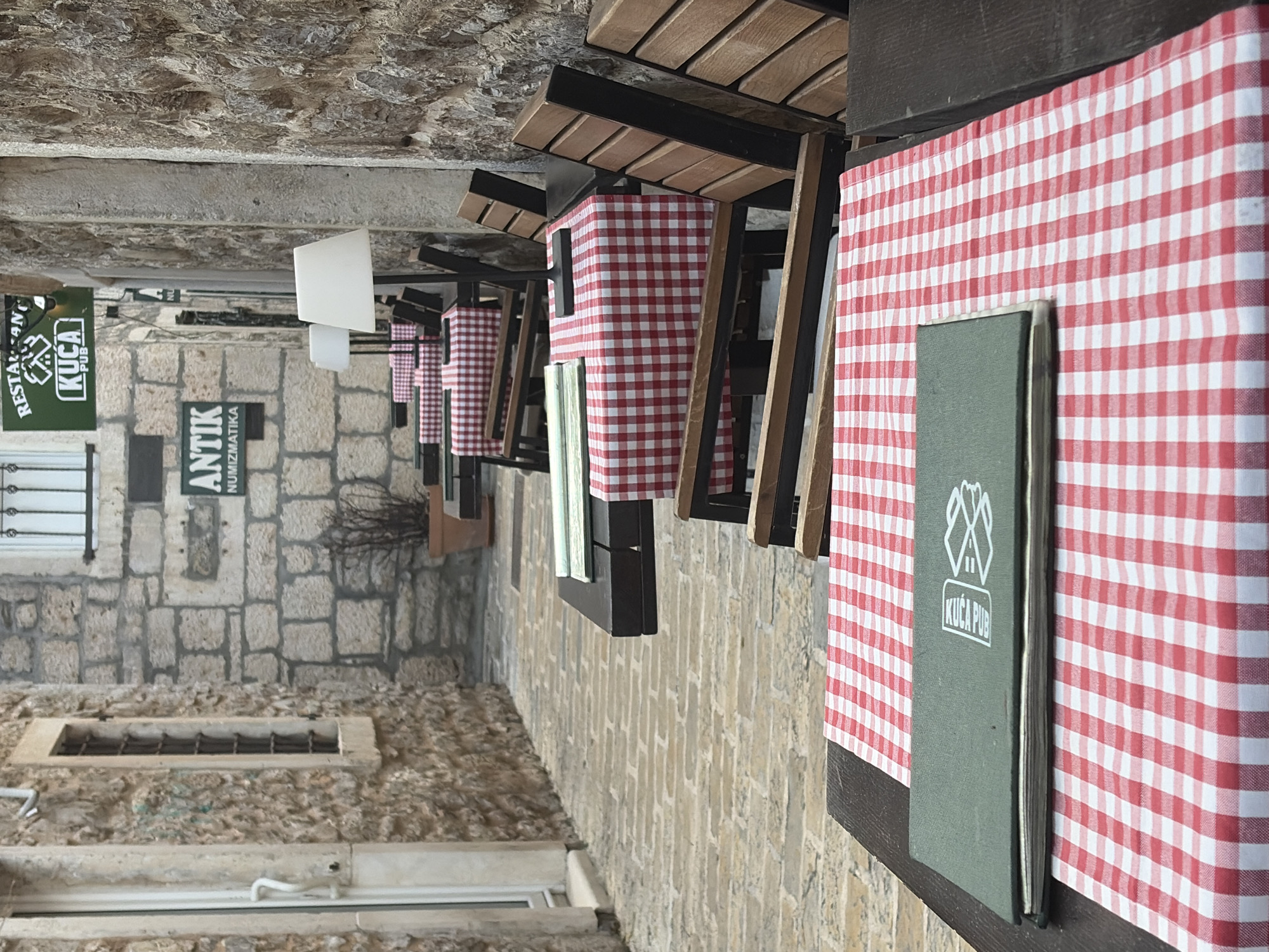 The image captures a scene in the city of Budva, Montenegro. It shows a row of small outdoor dining tables with red and white checkered tablecloths. Each table is set with a green menu bearing the logo "KUCA PUB". The tables are arranged along a narrow street with stone buildings on either side. One building has a sign saying "Restoran KUCA PUB" and another reads "ANTIK Numizmatika". There is a window with metal bars and some windowsills. The overall setting appears to be a restaurant or pub area within the old town.