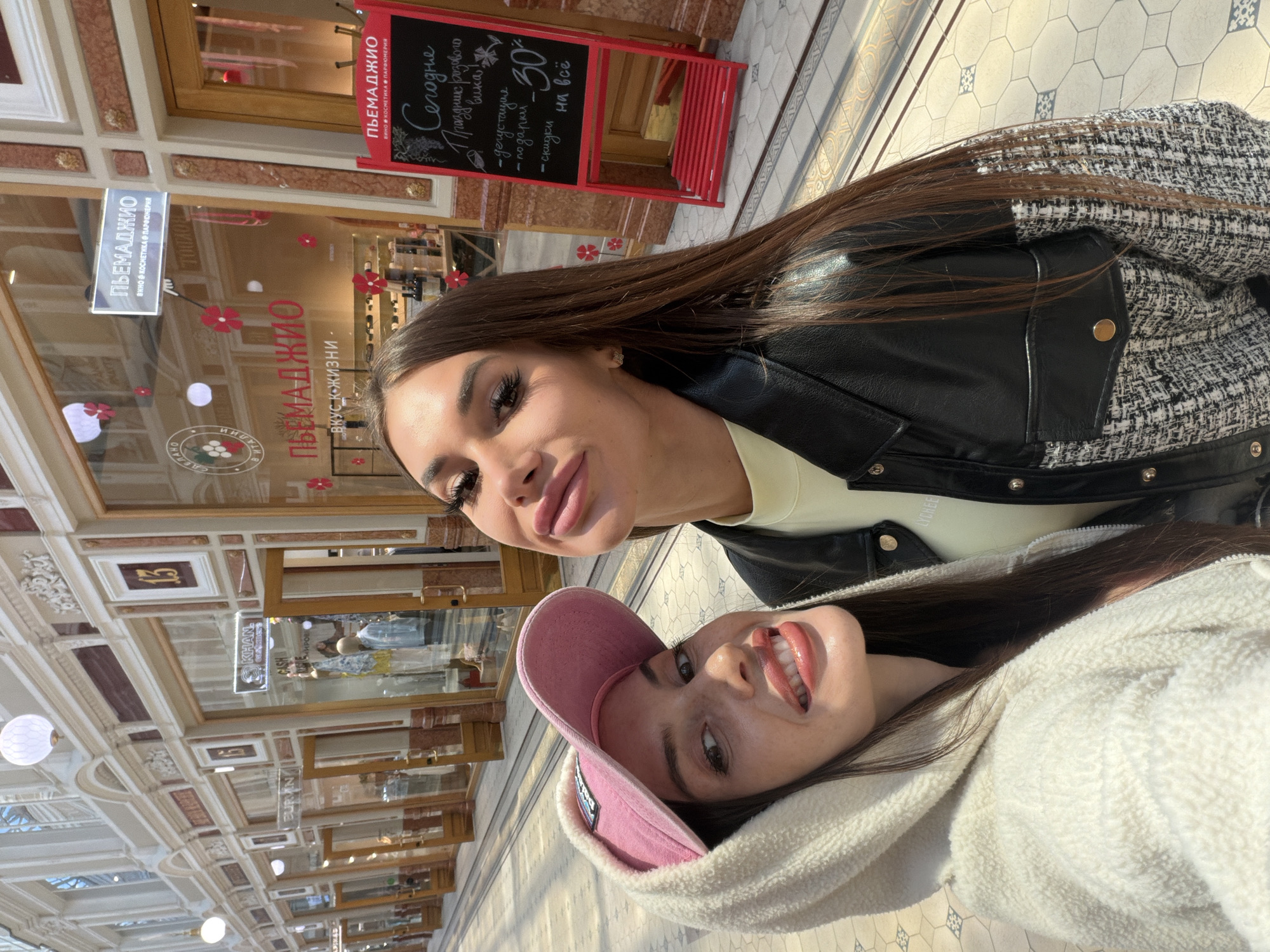 This media file is a selfie taken by two young women inside a brightly lit shopping arcade in Saint Petersburg, Russia. The women are positioned close together, with their faces filling the foreground. The woman closer to the camera is wearing a pink baseball cap and a cream-colored hooded jacket, and she is smiling widely, looking directly at the camera. Her makeup is prominent, with defined eyebrows and glossy lips. The second woman, positioned slightly behind and to the side, is also smiling, her face angled towards the camera. She has long, dark hair and is wearing a black leather jacket over a light-colored top.
In the background, the ornate architecture of the shopping arcade is visible, with arched ceilings, decorative plasterwork, and tiled flooring. Several shop windows and signs are present, indicating a commercial setting. One prominent sign reads "ПЬЕМАДЖИО" in large letters, with smaller text below it that translates to "wine, cosmetics, perfumery." Another sign, a red chalkboard with white lettering, advertises "Today - pink wine festival," with details about tasting, gifts, and discounts. The overall atmosphere appears cheerful and relaxed, with the sunlight casting patterns on the floor, suggesting it is daytime. The presence of the signs in Russian and the architectural style are strong cues for the location being Saint Petersburg.