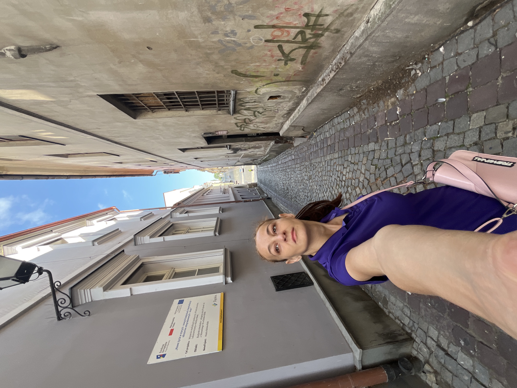 This is a selfie taken by a woman standing on a cobblestone street in a narrow alleyway. She is wearing a purple dress and a pink handbag is slung over her shoulder. The buildings on either side of the alley are made of brick and stone, with some showing signs of age and graffiti. The sky is blue and the weather appears to be sunny.
On the wall of one of the buildings, there is a sign with text in Polish. The visible text includes "Fundusze Europejskie" (European Funds) and mentions "Nowe życie w starych nowych budynkach" (New life in old new buildings). It also lists a project goal related to revitalization and an object described as a residential and service building. The sign also indicates "Miasto Bielsko-Biała" (City of Bielsko-Biała) as the beneficiary. This suggests the location is likely in Bielsko-Biała, Poland, and the photo was taken during the daytime. The woman's expression is neutral, and she is looking directly at the camera. The overall impression is a candid shot taken while exploring an urban environment.