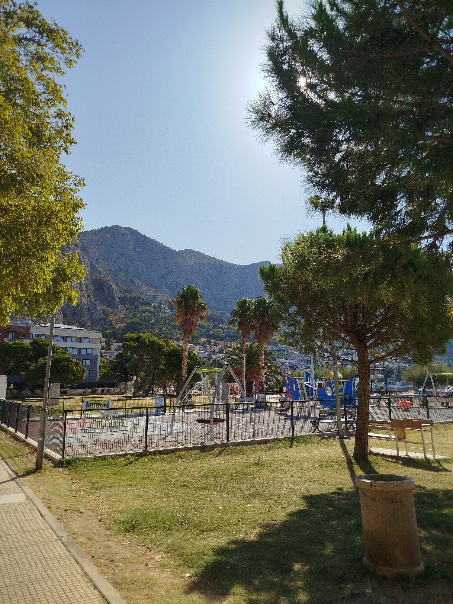 This is a daytime, eye-level, outdoor, full shot of a playground in Omiš, Croatia. The playground is fenced and contains various play structures, including swings and what appears to be a boat-themed climbing structure. A pathway made of light-colored interlocking pavers runs along the left side of the frame, adjacent to a grassy area. In the background, there are palm trees, other deciduous trees, and buildings, including a multi-story building with many windows. Beyond the buildings, a large, rocky mountain dominates the landscape under a clear blue sky. The sun is bright, casting shadows from the trees and playground equipment.