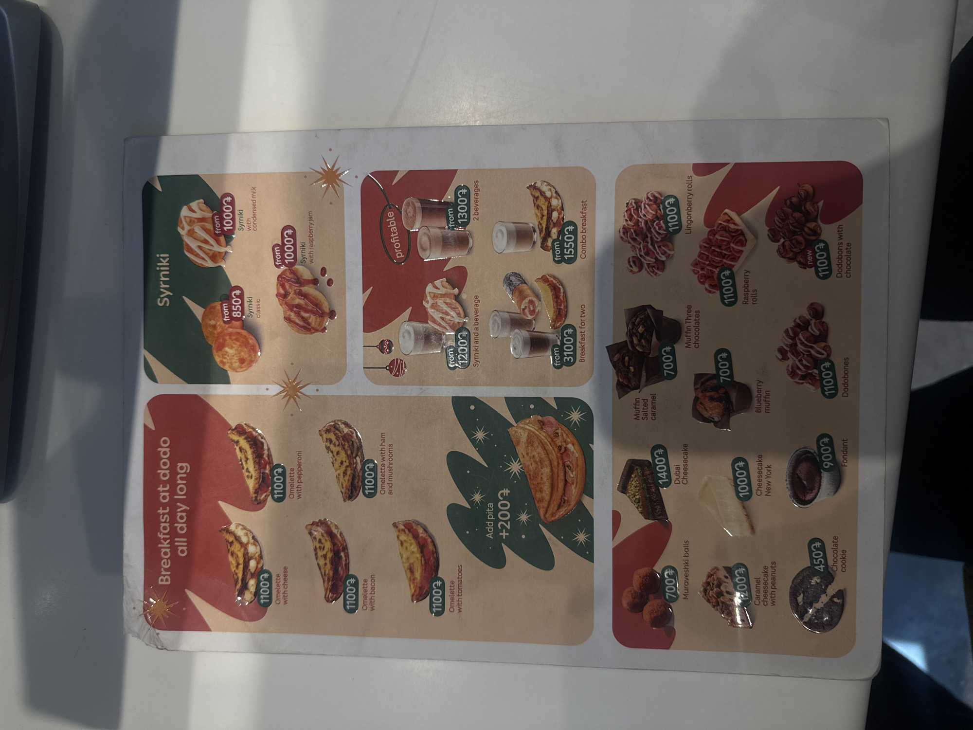 The image shows a close-up of a menu from Dodo Pizza in Yerevan, Armenia. The menu is printed on a white background with food items categorized as "Breakfast at dodo all day long" and "Syrniki". The breakfast items include omelets with various toppings like cheese, pepperoni, bacon, ham and mushrooms, and tomatoes. Syrniki options include classic, with condensed milk, and with raspberry jam. There are also combo options including beverages. Other items include Muraveshki balls, cheesecakes, muffins, rolls, Dodobones and cookies. Prices are listed next to each item in an unspecified currency, denoted by a symbol resembling "7".