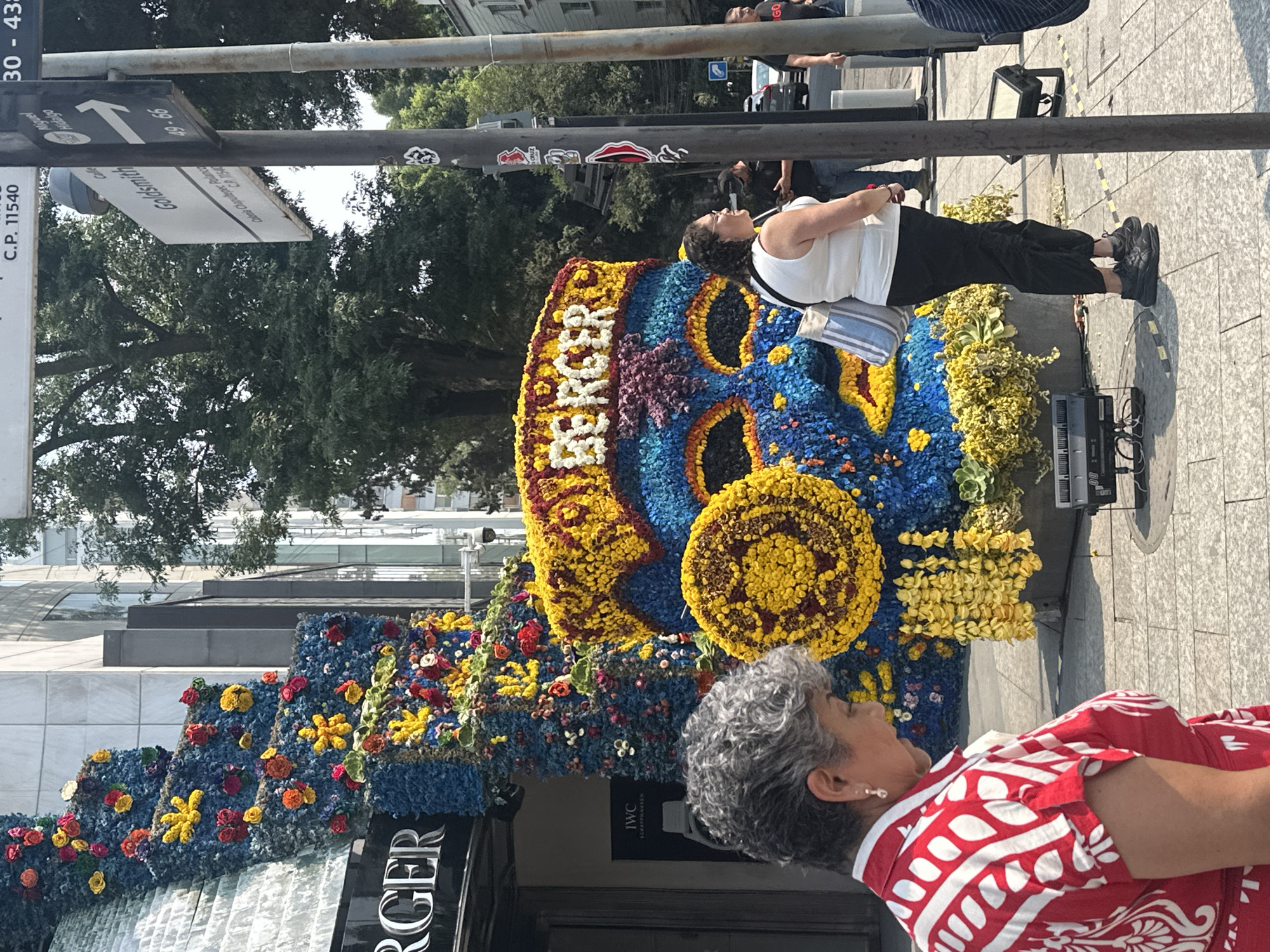 Here is a description of the image:
The image shows an outdoor scene, likely in Mexico City, Mexico. A large floral display is prominent, reading "BERGER". An IWC advertisement is visible nearby. Two women are present, seemingly observing the display. The environment appears to be a city street during the daytime, with a street sign indicating "Goldsmith" and a street number "C.P. 11540" suggesting the Polanco neighborhood. The scene appears to be a daytime event or installation on a city street.