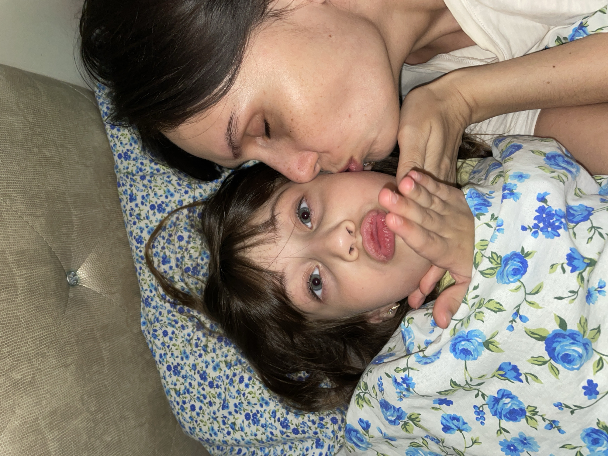 A woman with dark hair kisses a young child with dark hair and brown eyes. They are lying down on a bed with a blue floral-patterned blanket. The background shows a portion of a tan-colored headboard. The location is unknown.