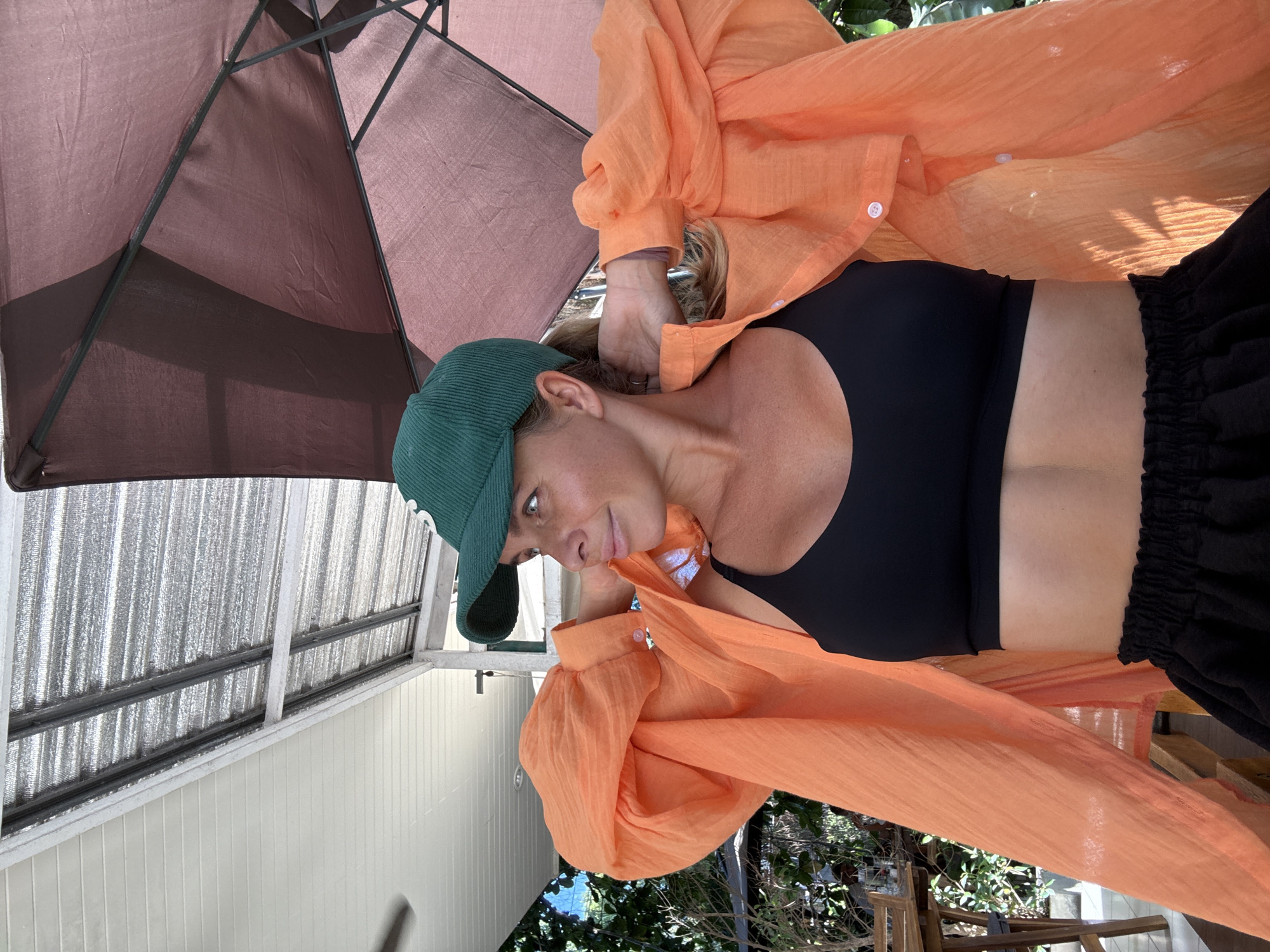 A woman wearing a green baseball cap, an orange unbuttoned shirt, and a black sports bra poses for a photo under a brown umbrella. The woman is looking to her left with her hands behind her head. She is outdoors in Ko Samui District, Thailand, with a thatched roof structure and trees visible in the background.