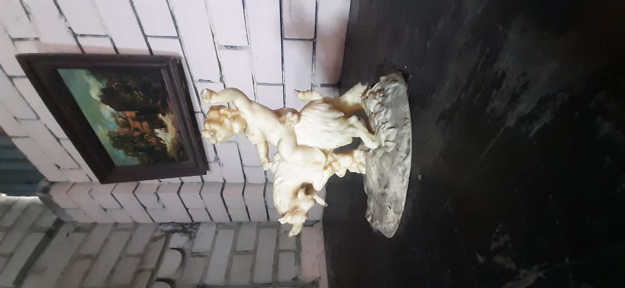 A light-colored ceramic figurine of a cherubic child riding a goat is displayed on a dark, polished surface. The child is depicted in motion, with one arm raised. The figurine rests on a textured, uneven base. In the background, a framed landscape painting hangs on a white brick wall. The painting shows a rustic scene with trees and buildings under a cloudy sky. The scene is lit from above, casting shadows on the subjects and surface. The location is in Yessentukskaya, Russia.