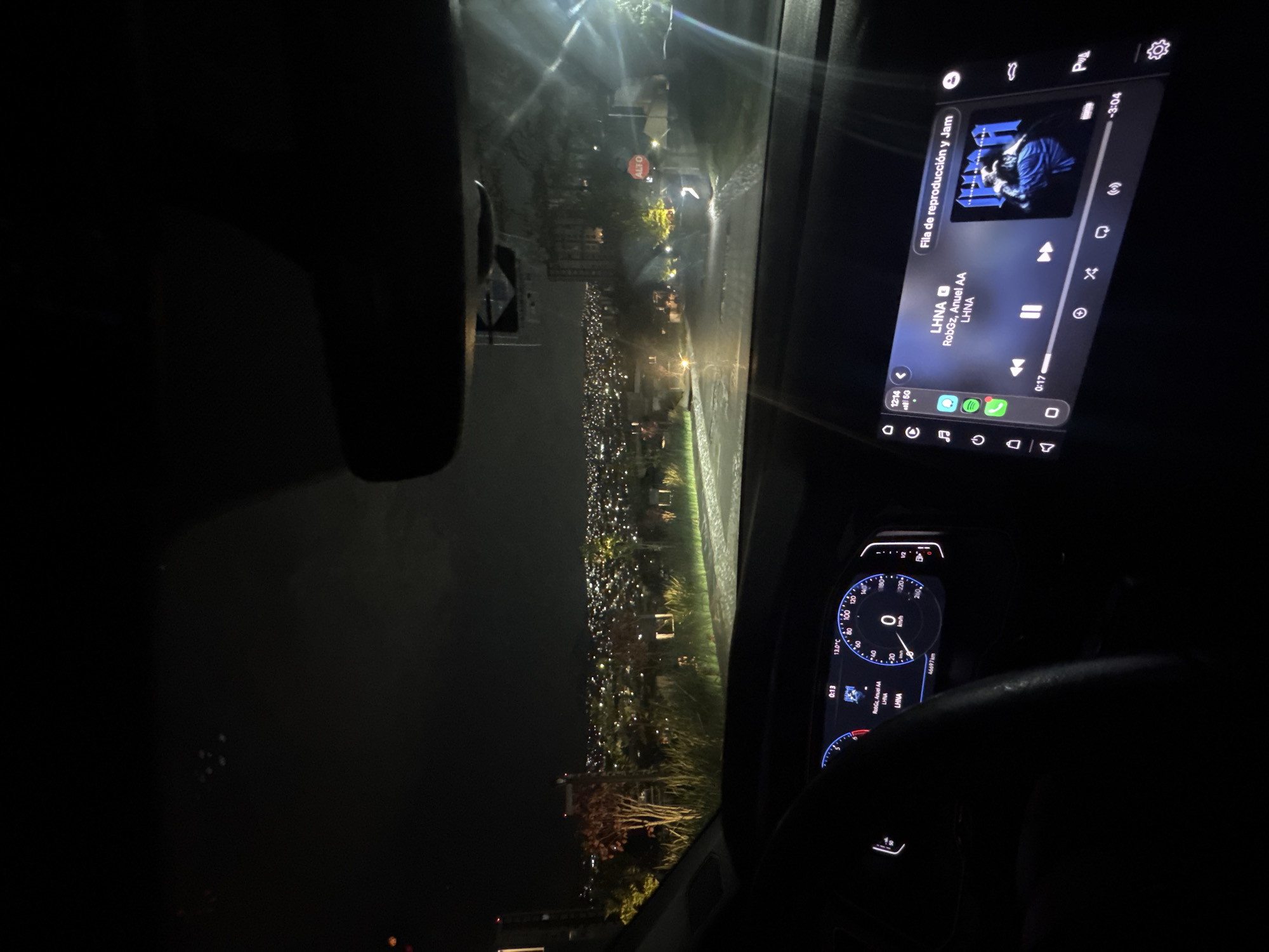 Here's a description of the image:
The image is taken from inside a car driving at night in Huixquilucan de Degollado, Mexico. The car's infotainment screen displays a song titled "LHNA" by RobGz and Anuel AA, and the odometer shows 46697 km. The speedometer indicates a speed of 0 km/h, and the temperature is 13.0°C. Through the windshield, a view of the city illuminated at night with lights can be seen. An "ALTO" (Stop) sign is visible on the road ahead.