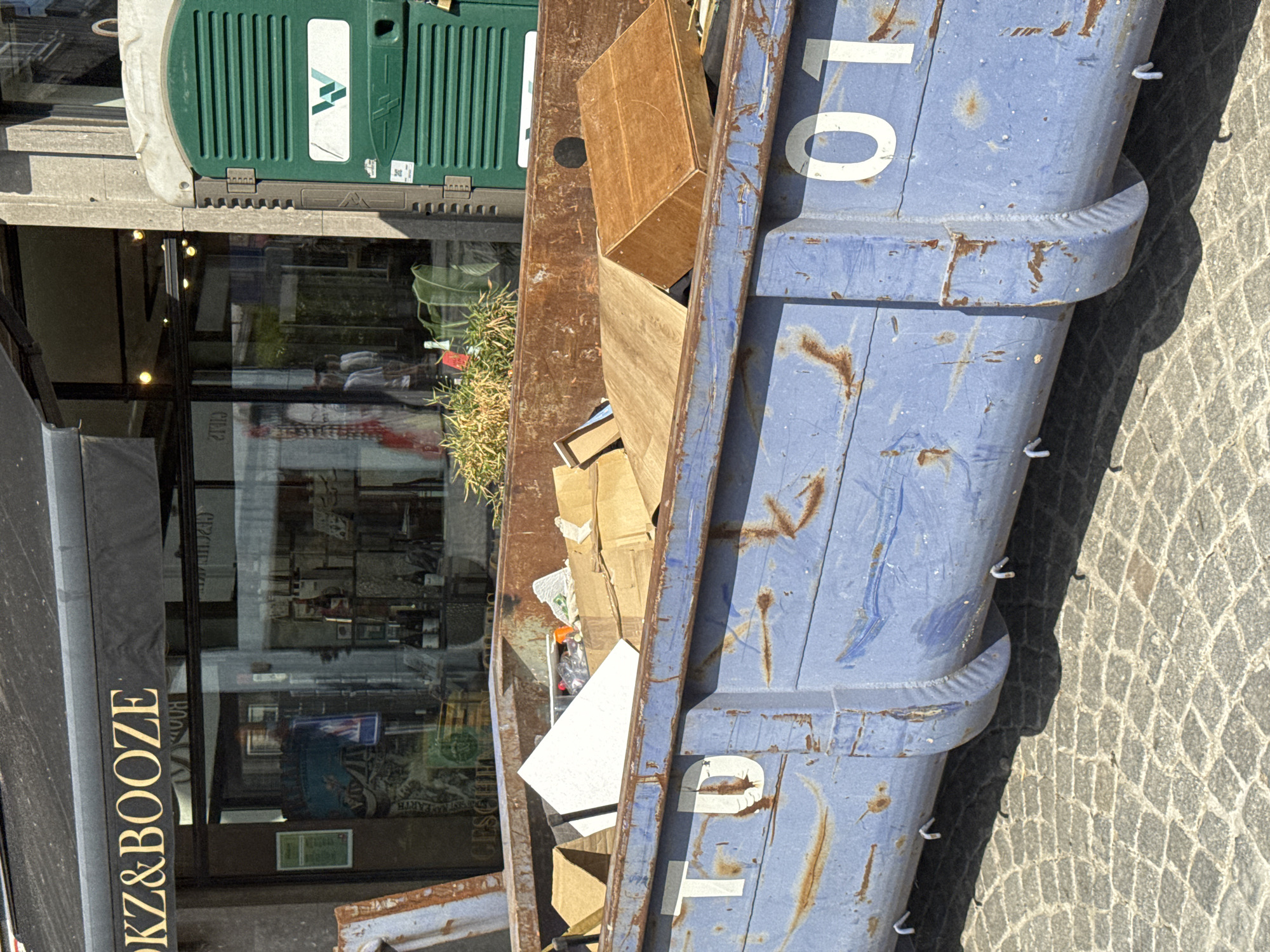 An outdoor, eye-level, medium shot shows a large, blue, rusted dumpster filled with debris. The dumpster is positioned on a cobblestone street in front of a building with a black awning and glass windows. The awning displays gold lettering that reads "OKZ&BOOZE". Inside the shop, shelves are stocked with bottles and other items, and several mannequins are visible. To the right of the dumpster is a green portable toilet with a white logo depicting a green "W" shape. The dumpster has white numbers painted on its sides; "T" and "D" are visible on the left side, and "01" is visible on the right. Plants are growing in front of the shop, near the dumpster. The scene appears to be in an urban environment. The location is the city of Antwerp, Belgium.
