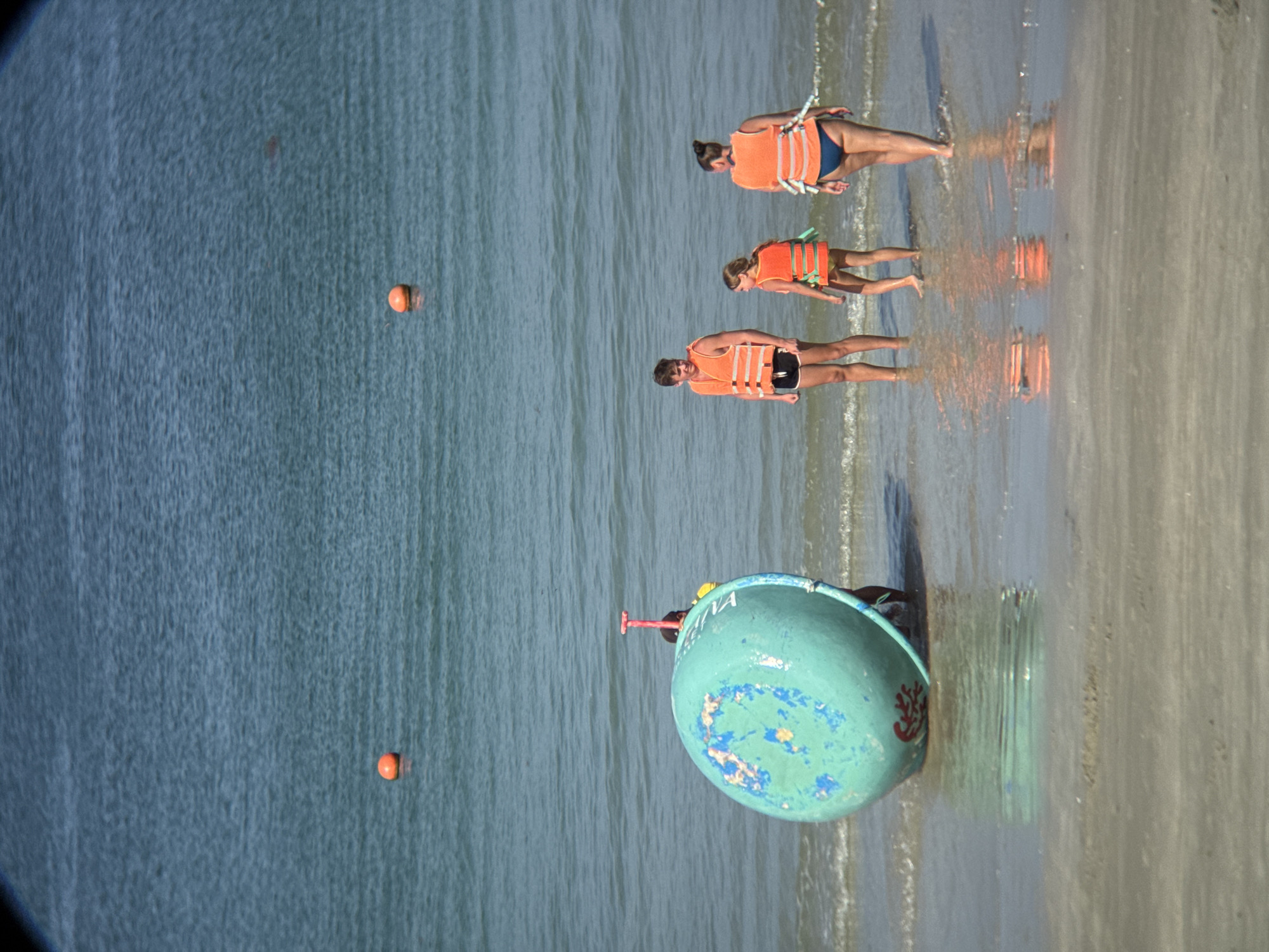 A group of three individuals, two adults and one child, stand in shallow water on a sandy beach. All three are wearing bright orange life vests. The adult male on the left is wearing black shorts. The adult female on the right is wearing a blue swimsuit and her hair is pulled back. The child, between them, is wearing a yellow swimsuit and a green object is visible around their waist. In the foreground, to the left, is a large, teal-colored cylindrical object with blue and white markings, possibly a buoy or a container. The ocean is calm and blue, with a few orange buoys visible in the distance. The scene appears to be in Đông Ninh Hòa, Vietnam, a coastal town known for its beaches and proximity to Cam Ranh Bay.