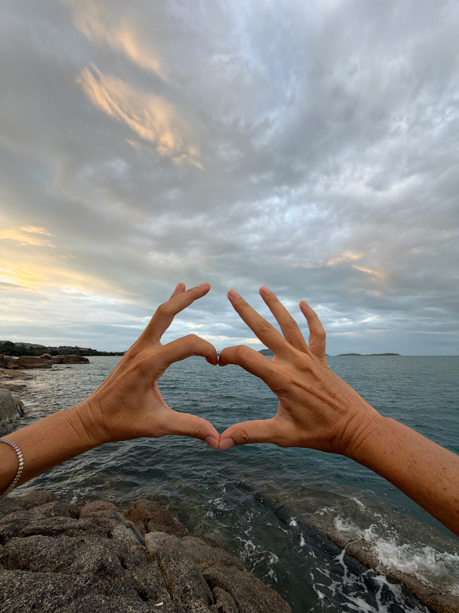 The image shows two hands forming a heart shape against a cloudy sky and a body of water. The hands are positioned in the foreground, with the thumbs and index fingers touching to create the heart shape. The background features a dark, choppy sea meeting a rocky shoreline. In the distance, a landmass with some vegetation is visible, likely part of the coast of Ko Samui. The sky is filled with dramatic, grey clouds, with hints of warm light breaking through on the left side, suggesting either sunrise or sunset. The overall impression is of a serene, natural landscape with a human gesture of affection.