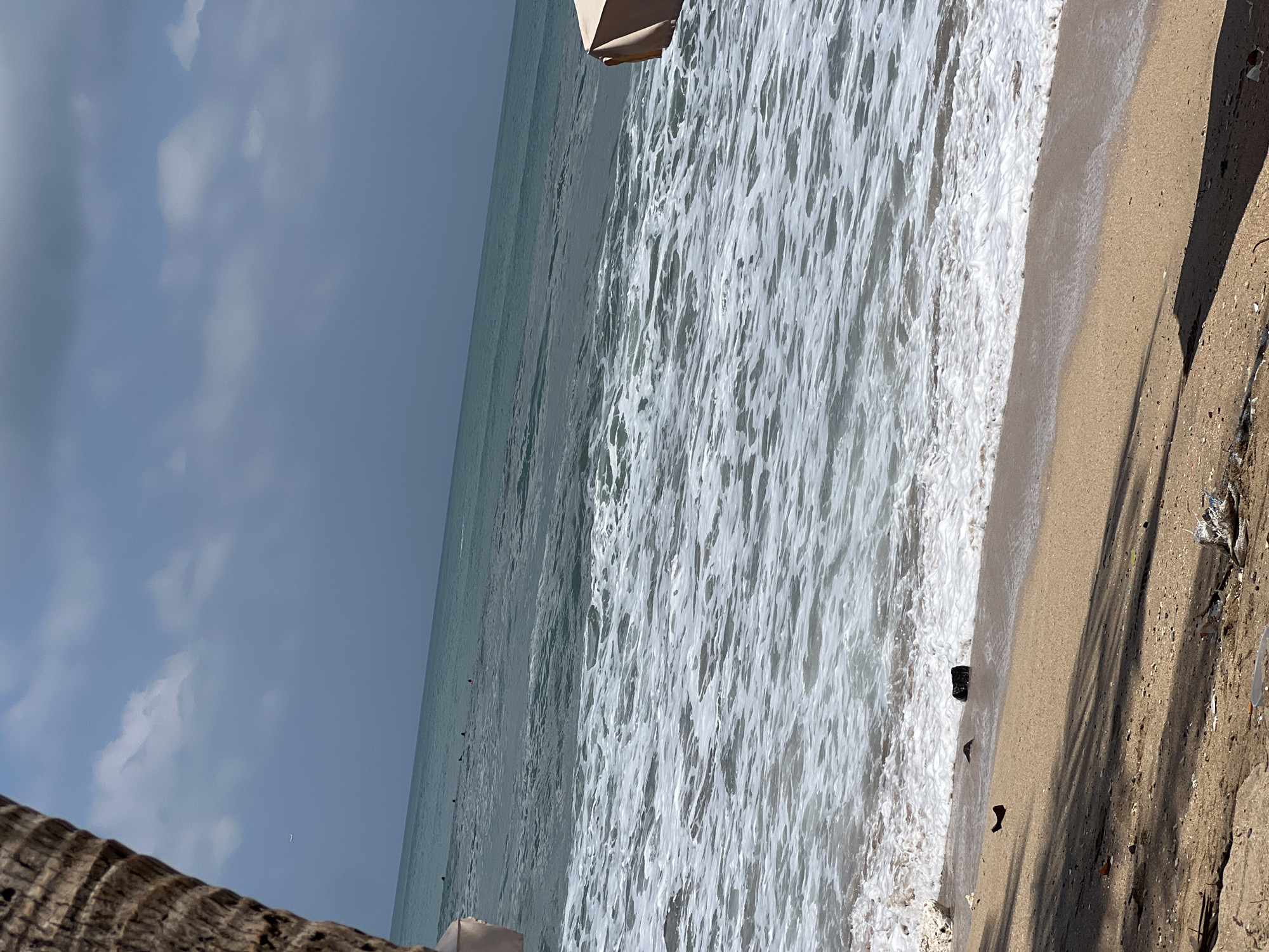 The image depicts a beach scene in Kuta, Indonesia. In the foreground, the sandy beach is littered with debris, including plastic waste and what appears to be wood fragments. A shadow from palm fronds or similar vegetation stretches across the sand. The ocean in the middle ground is characterized by its turquoise-green hue and white foamy waves crashing onto the shore. Several dark shapes, possibly surfers or swimmers, are visible in the water further out. The horizon is marked by the meeting of the sea and a hazy, overcast sky with scattered white clouds. A portion of a palm tree trunk is visible on the far left, and the edge of a beach umbrella is seen on the right. A faint white line, potentially a distant airplane contrail, is visible in the upper left quadrant of the sky.