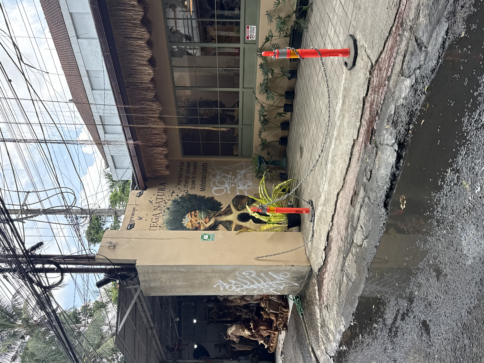 The image shows a street scene in Ubud District, Indonesia. In the foreground, there is a section of damaged pavement with a large puddle. Adjacent to the pavement is a sidewalk constructed of stone tiles. Two orange traffic cones are connected by a chain. Behind the cones, there is a building facade. The facade is beige and features a mural of a person with dark curly hair and a mask. The mural includes the text "EGA NATURA" and a quote attributed to Mahatma Gandhi: "You must be the change you want to see in the world." Below the mural, there is some graffiti and an open area filled with wooden carvings. Above the mural, the building has windows with green frames and a thatched roof overhang. In the upper left of the image, there are numerous tangled wires overhead. The sky is visible in the background. The overall scene appears to be in a commercial or tourist area, showing some signs of urban decay.