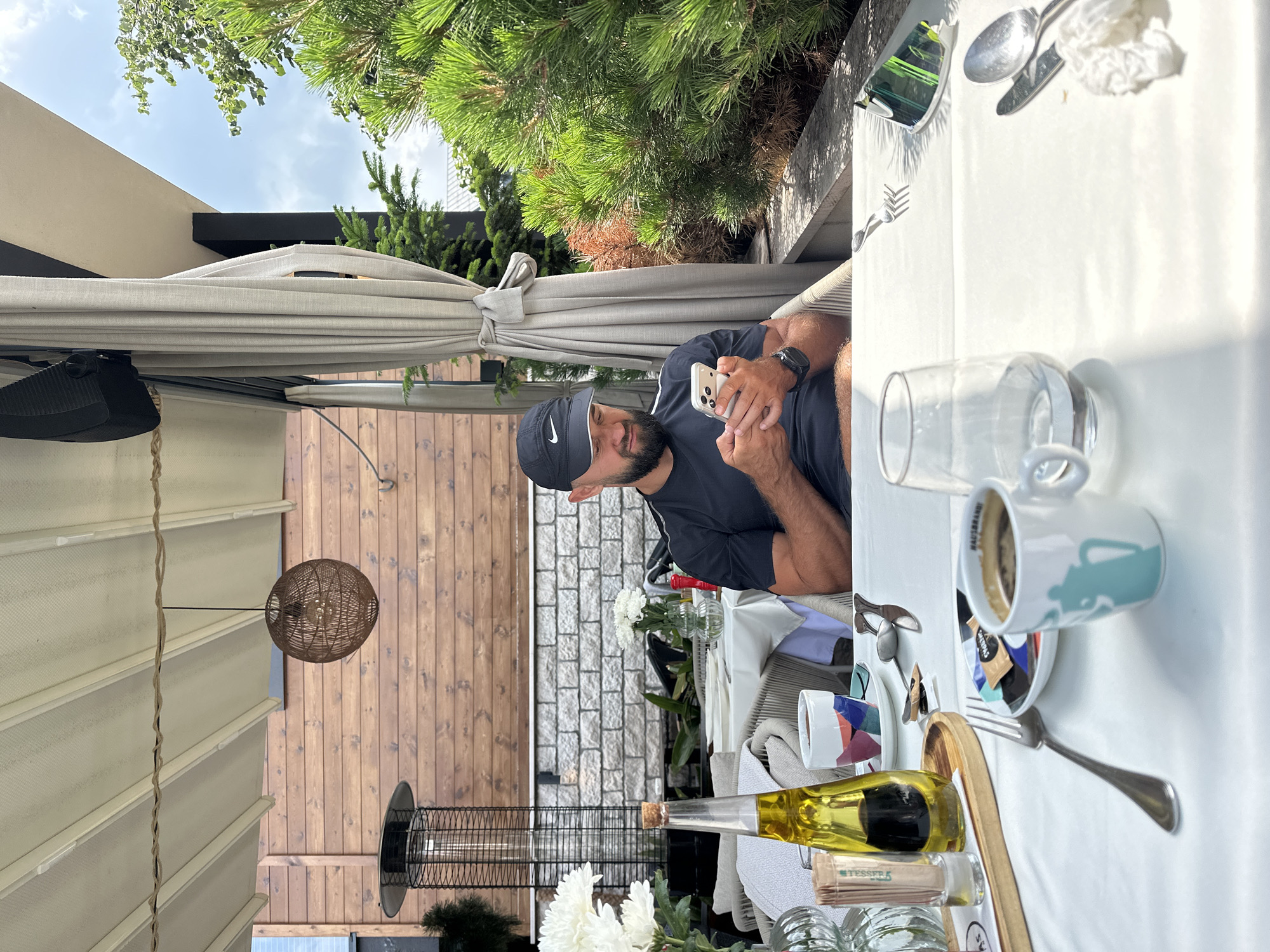 A man with a beard and a black Nike baseball cap is sitting at a table outside, likely on a patio or terrace, and is looking down at his smartphone. He is wearing a black t-shirt. The table is covered with a white tablecloth and set with a coffee cup, a small plate with sugar packets, a fork, a knife, and a spoon. A bottle of olive oil and a glass of water are also on the table. In the background, there is a wooden wall, a patio heater, and some greenery. The location appears to be a cafe or restaurant. It is daytime and sunny. The setting suggests a casual outdoor dining experience in a city, possibly Kolašin, Montenegro, based on the provided context.