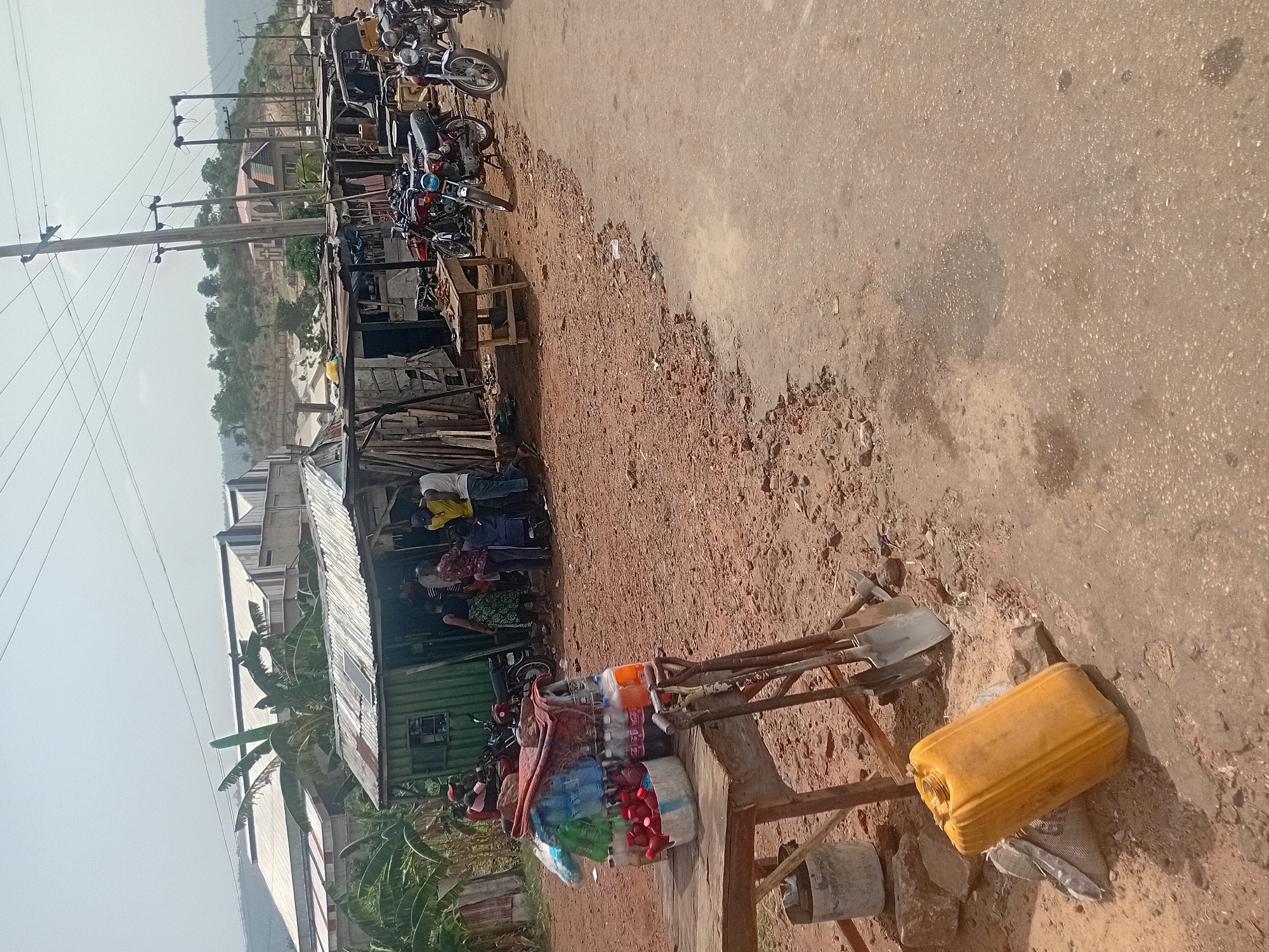 The image depicts a bustling roadside scene, likely a market or commercial area, under bright daylight. The ground is a mix of reddish-brown dirt and a paved, light-colored road surface, with visible erosion where the two meet.
In the immediate foreground, on the dirt section, a wooden stand holds several shovels and what appears to be a bucket. Nearby, a yellow plastic jerrycan rests on the ground. A makeshift roadside stall, constructed from corrugated metal sheets and possibly wood, dominates the left side of the frame. This stall displays various goods, including numerous plastic bottles, some in a white basket, suggesting beverages or water for sale.
Several individuals, mostly adults, are visible around the stall, some appearing to be customers or vendors. A cluster of motorcycles is parked on the dirt area next to the stall, indicating a common mode of transport.
Beyond the immediate foreground, a series of utility poles with power lines extend into the distance, running parallel to the road. Further back, various buildings with different architectural styles and roof types are visible, interspersed with green vegetation, including large-leaved plants that resemble banana trees. In the farthest background, rolling hills or mountains are silhouetted against a bright, possibly hazy, sky.
The scene portrays daily life and commerce in what appears to be a semi-urban or rural setting. No discernible text is visible in the image.
