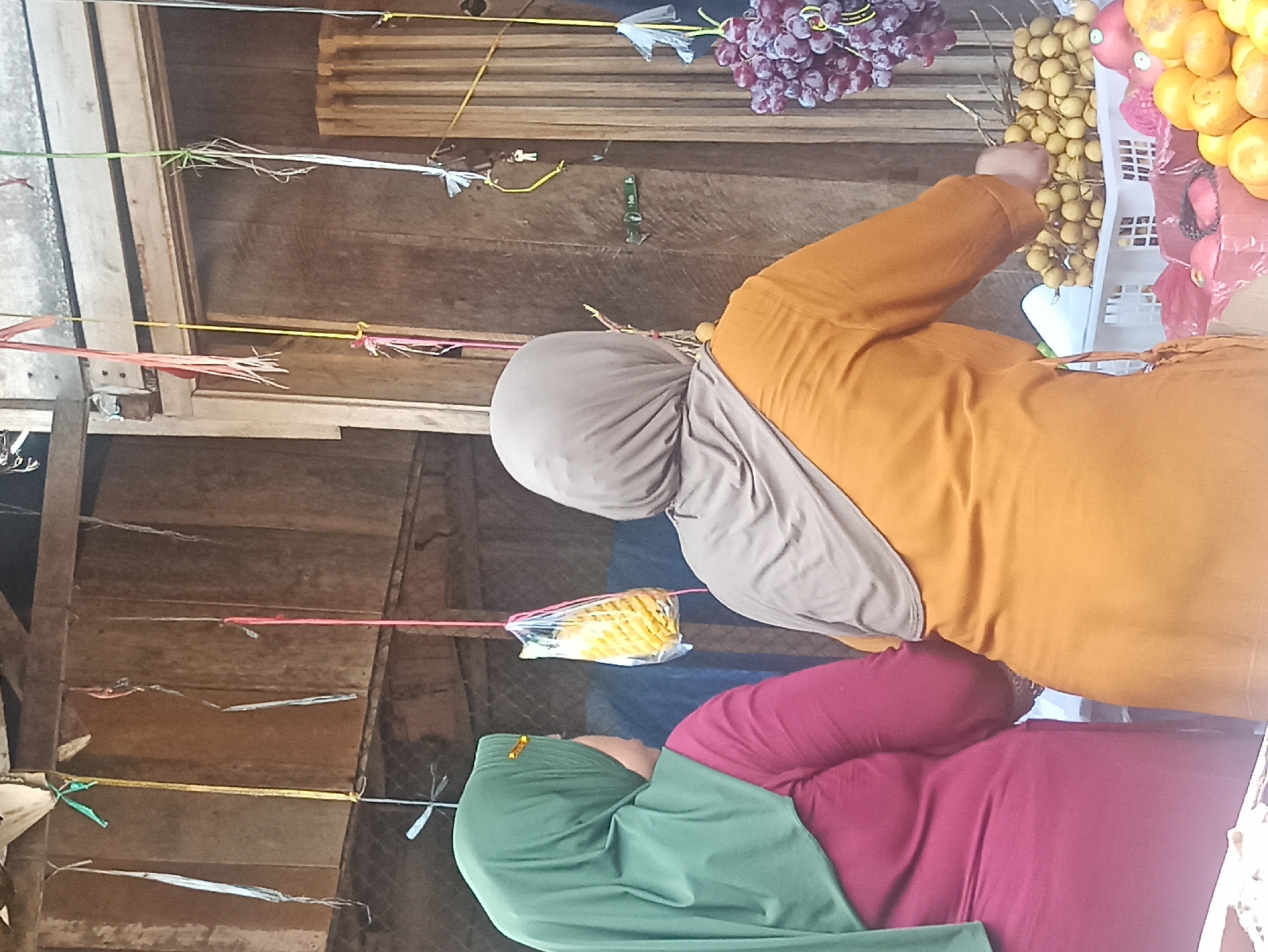 In Tuba, Indonesia, two women wearing hijabs are browsing a fruit stall. The woman on the left wears a green hijab and a magenta top, while the woman on the right wears a beige hijab and an orange top. They are positioned with their backs to the camera, facing a display of various fruits. On the stall, visible fruits include bunches of purple grapes, longan, and what appear to be oranges and pomegranates. The stall is constructed of wood, and the scene is lit by overhead lighting, with various strings and decorative items hanging from the ceiling. A plastic-wrapped pineapple is also visible hanging from one of the strings.