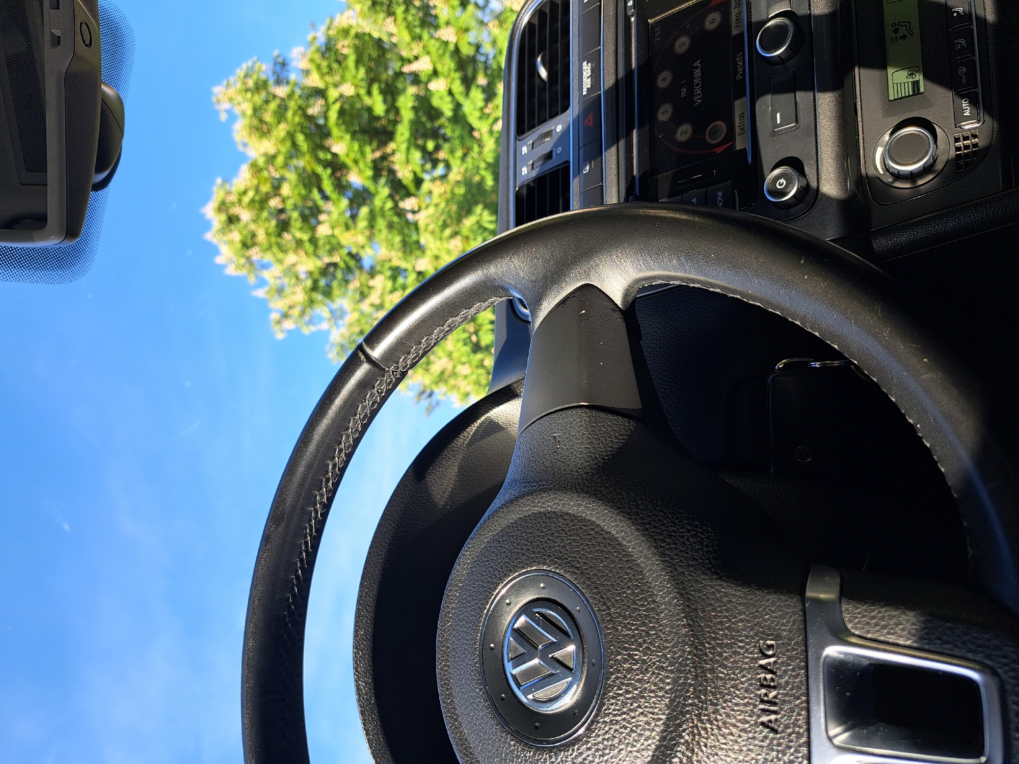 The photograph is taken from inside a car, focusing on the steering wheel which features a prominent Volkswagen logo. The dashboard and infotainment system are partially visible to the right of the steering wheel, along with climate control buttons. The view through the windshield shows a blue sky and the green foliage of a tree. A rearview mirror is visible. The location in the city of Sofia, Bulgaria cannot be ascertained from the image.