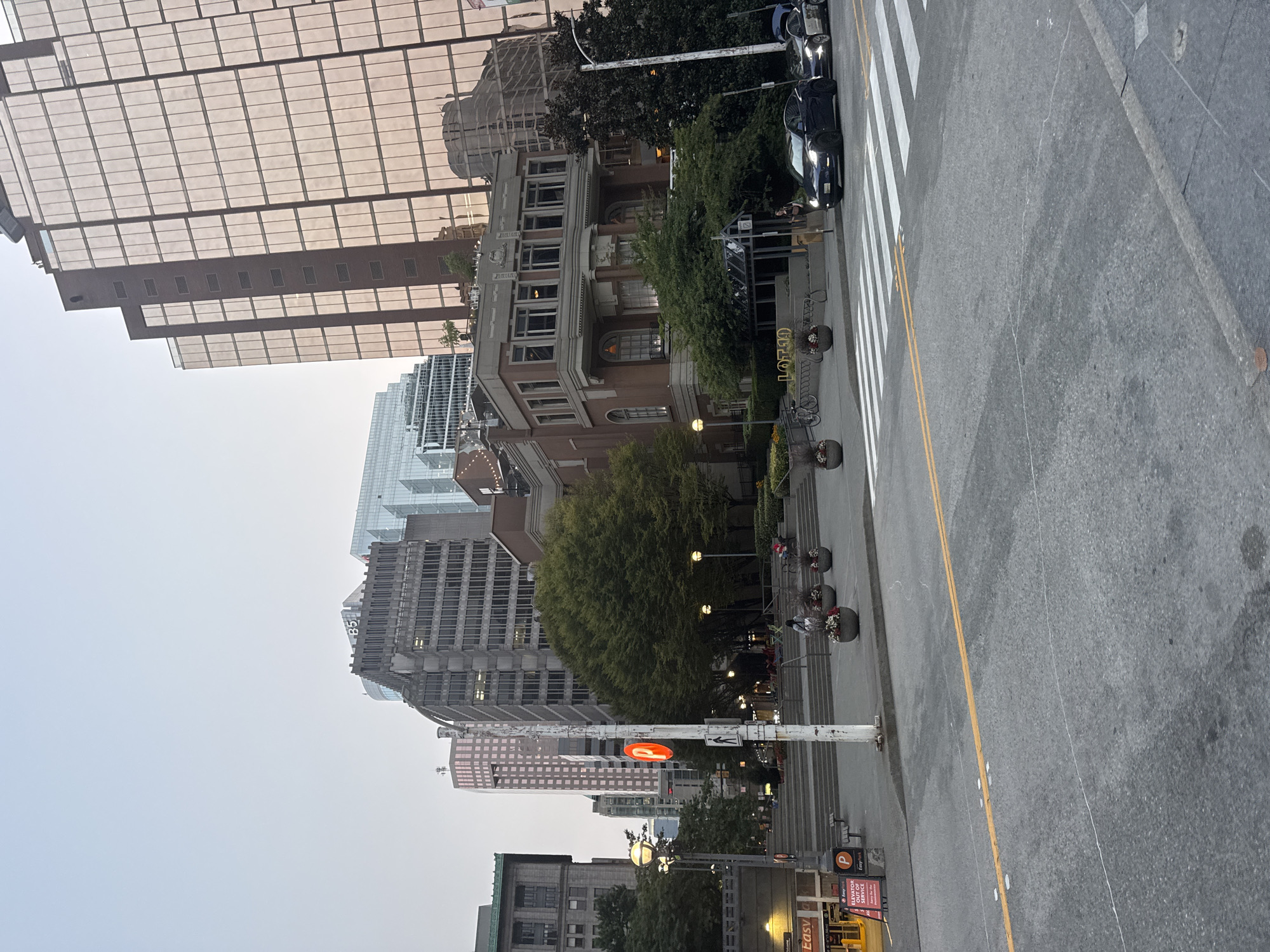 The image captures a street scene in Vancouver, Canada, likely in the downtown core. The perspective is from street level, looking up towards a collection of buildings and an outdoor public space. Dominating the background are several tall, modern buildings with glass facades, reflecting the overcast sky. One prominent building features a rose-gold tinted glass. In the mid-ground, a classical-style building with red brick and ornate details stands out, with the word "LOT3D" visible in large letters above its entrance.
In the foreground, a paved street with a pedestrian crosswalk is visible, with yellow lines marking the road. Two dark-colored cars, possibly Teslas, are stopped at the crosswalk. To the left of the street, a set of concrete stairs leads up to a plaza area. Several large planters filled with flowers are positioned on the plaza. A white lamppost with a red "P" sign for parking is also visible. The environment appears to be a busy urban area, with elements of both modern and older architecture. The lighting suggests it is either early morning or late afternoon, with diffused natural light. There are a few individuals visible in the scene, some walking on the plaza and others in or near the cars, but their actions are not clearly discernible. The overall impression is a city intersection with a mix of commercial and public spaces.