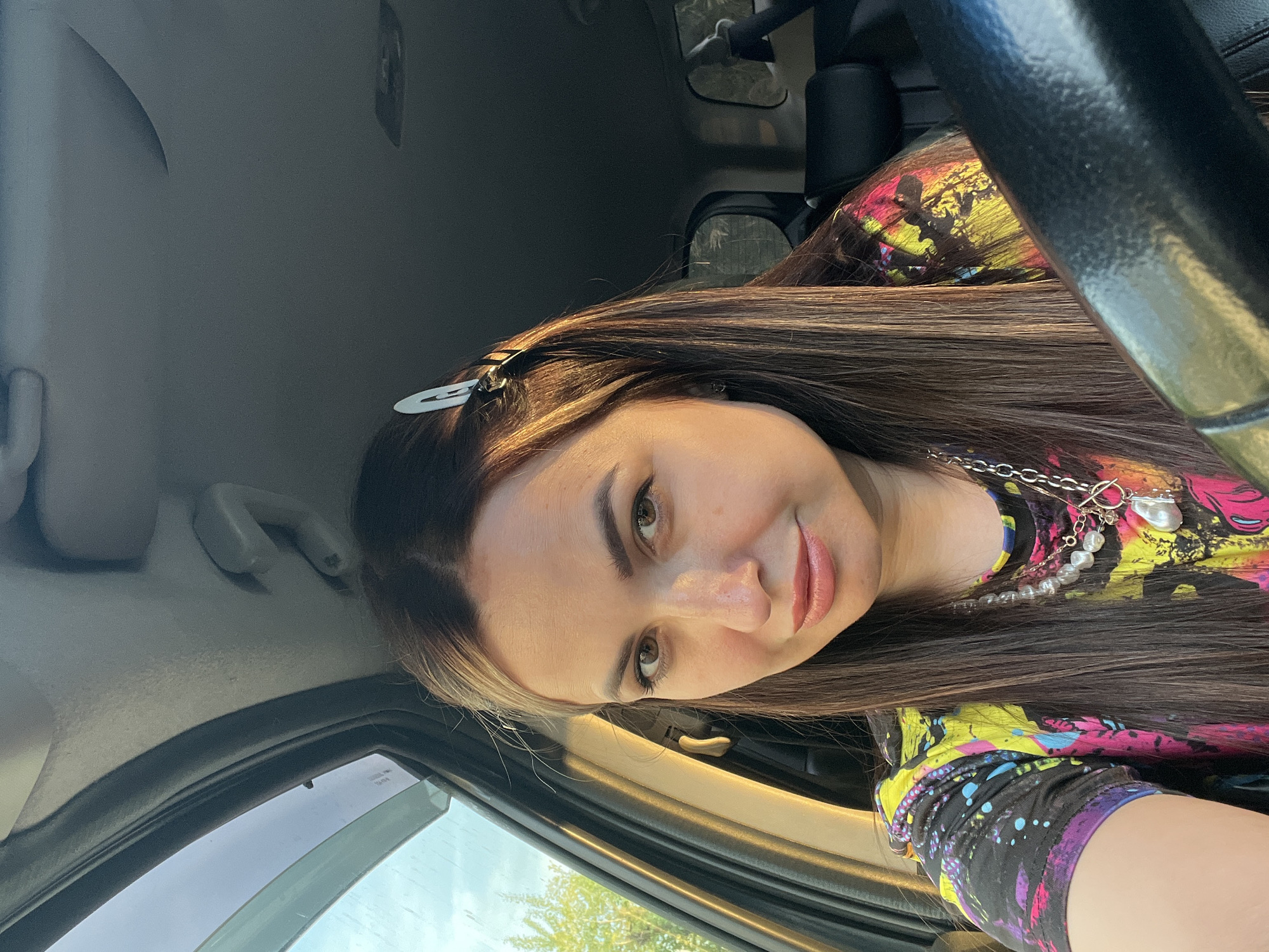 A woman with long brown hair is pictured from a close-up angle, likely seated inside a vehicle. She is wearing a brightly patterned top and a necklace with a silver chain and pearls. Her hair is adorned with a silver clip. The interior of the car is visible, including the steering wheel, dashboard, and a glimpse through the window showing some greenery. The location appears to be a city; however, the specific location remains Unknown, Unknown.