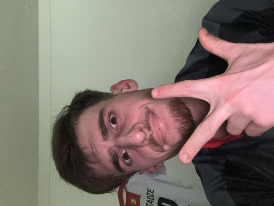 A man with brown hair and a trimmed beard poses, making a "V" sign with his fingers near his face. He looks directly at the camera with a slight smirk. He wears what appears to be a jacket and a white shirt with text that reads "Stadze" on it. The background is a plain wall, possibly indoors. The image appears to be taken at an odd angle, rotated slightly.